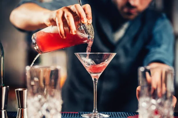 Bartender pouring red cocktail into a martini glass at a bar.