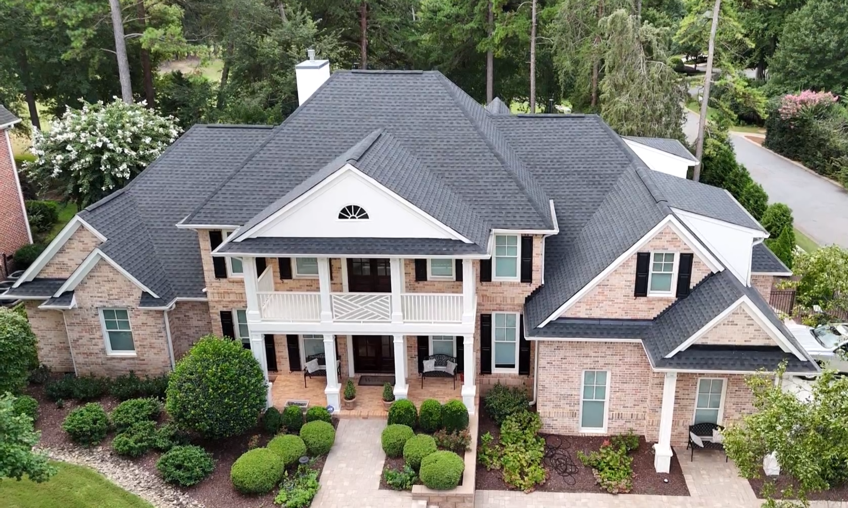 A large, two-story brick house with a new charcoal shingled roof. The front yard is landscaped with bushes, shrubs, and trees, with a stone pathway leading to the front entrance.