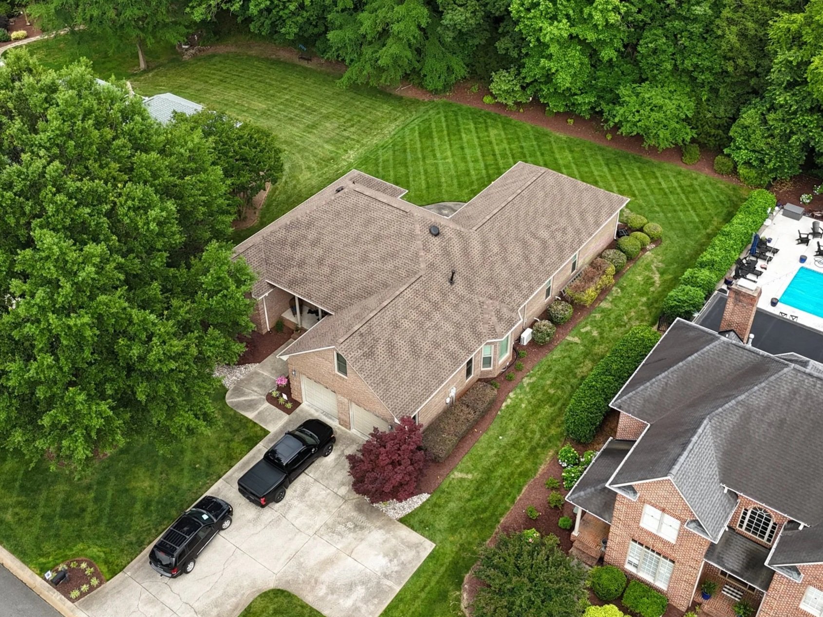 An aerial view of a suburban backyard featuring a single-story brick house with a new brown shingle roof, a driveway with three black cars, a large green lawn, trees.