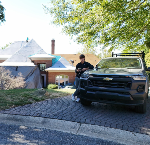 Felipe standing next to a work truck on a driveway in front of a house with a steep roof and chimney, undergoing a roof replacement