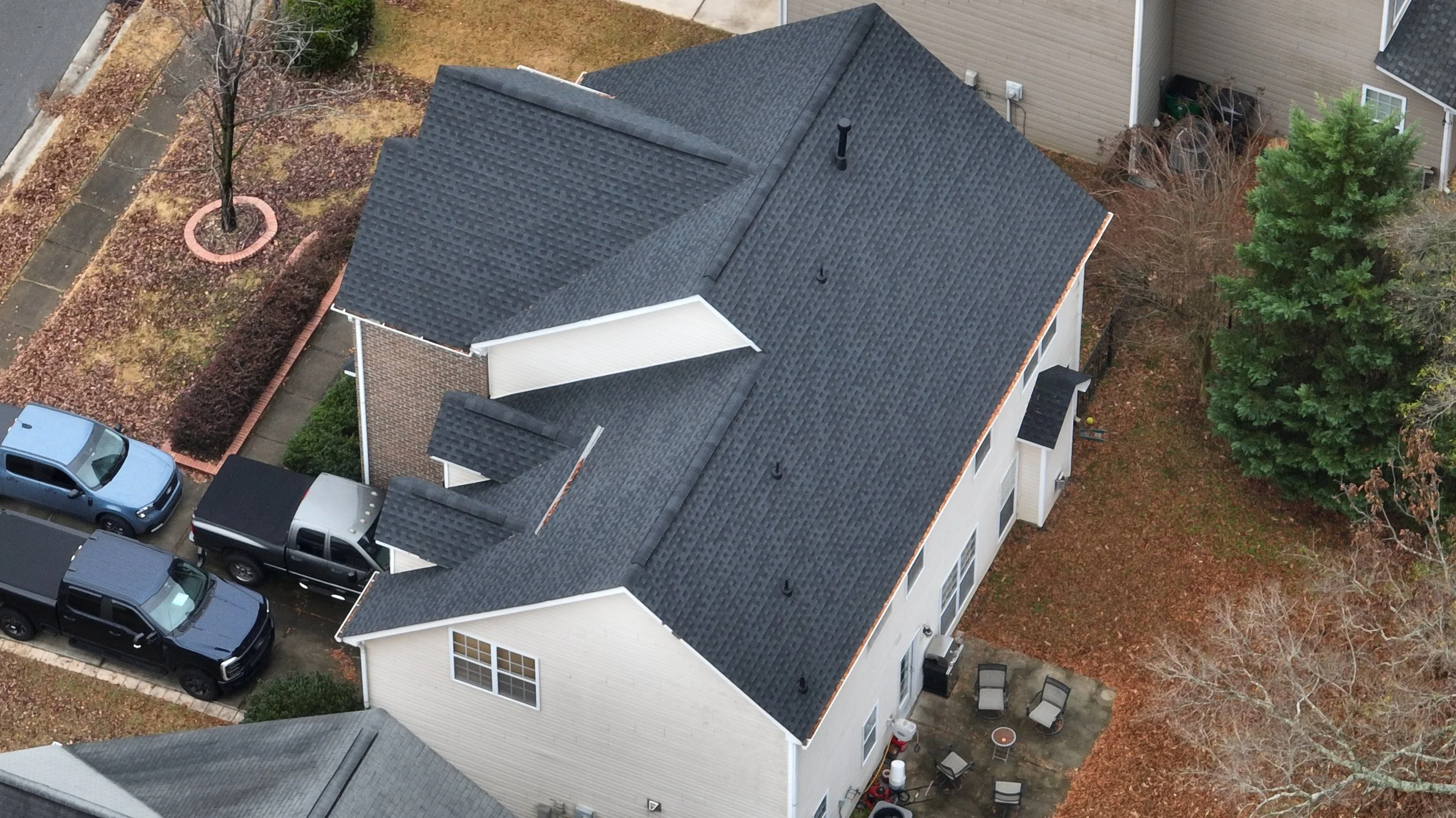 An aerial view of a house with a dark gray shingle roof, parked cars, and surrounding trees with fallen leaves.