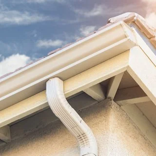Close-up of a house's roof corner showing a beige downspout attached to the gutter and house wall.