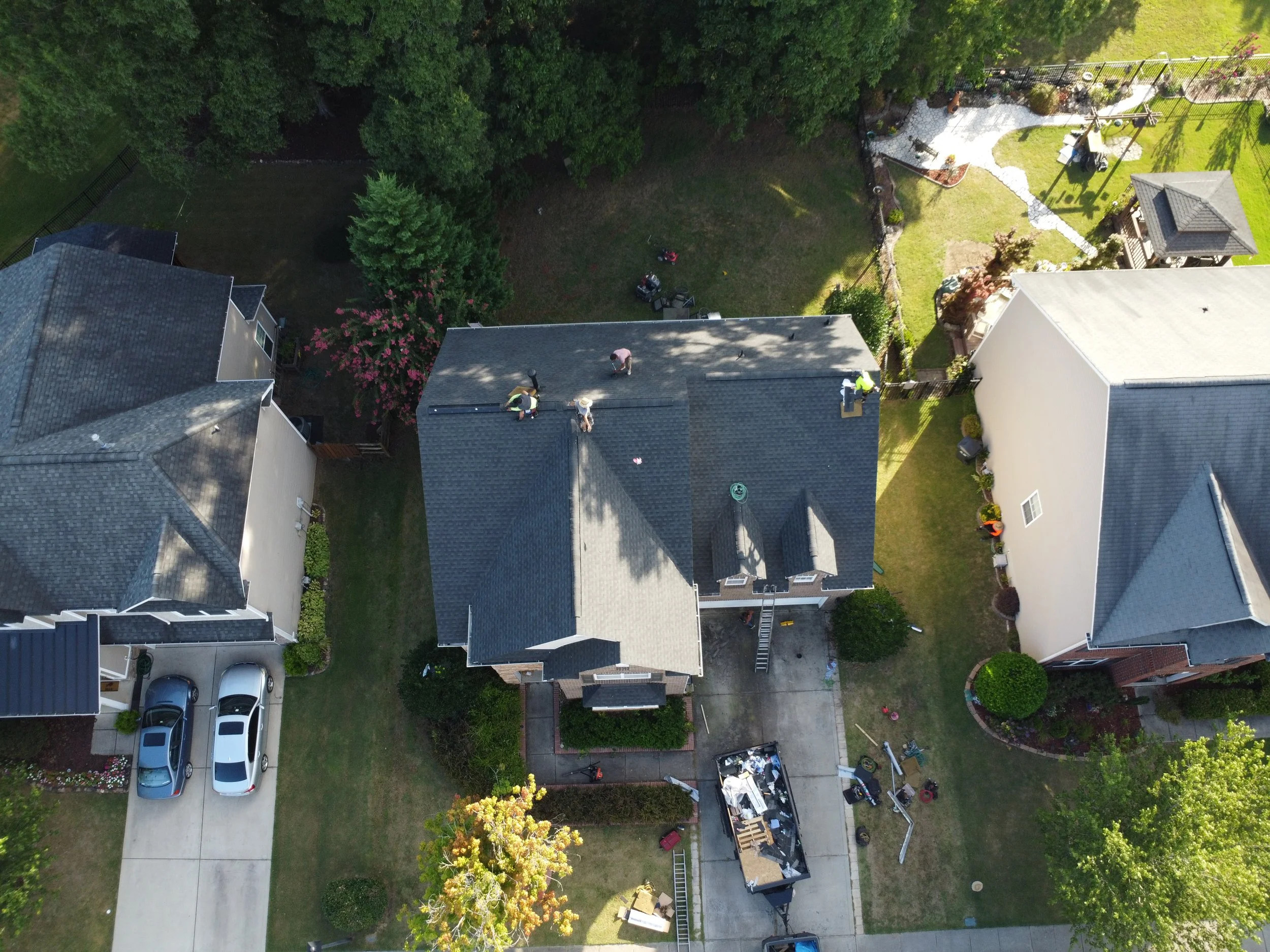 Aerial view of a residential neighborhood showing a house under roof repair with workers on the roof, neighboring houses, a driveway with cars, and a backyard with trees and a garden.
