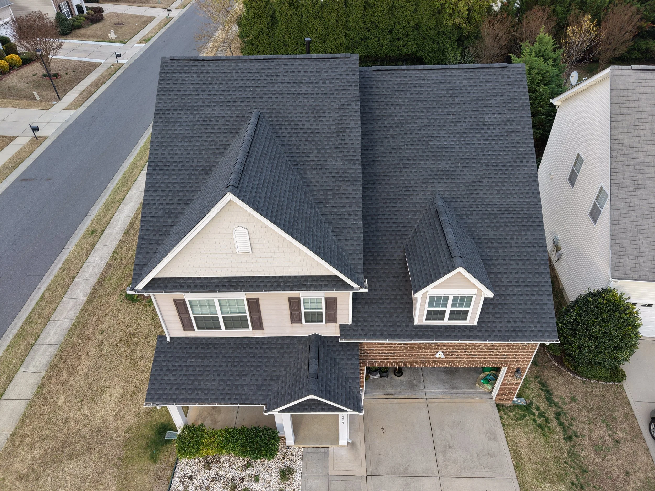 An aerial view of a two-story house with a black shingled roof, beige siding, and a brick facade on the front. There is a covered porch at the entrance and a two-car garage. The house is surrounded by a small lawn and neighboring houses and streets are visible.