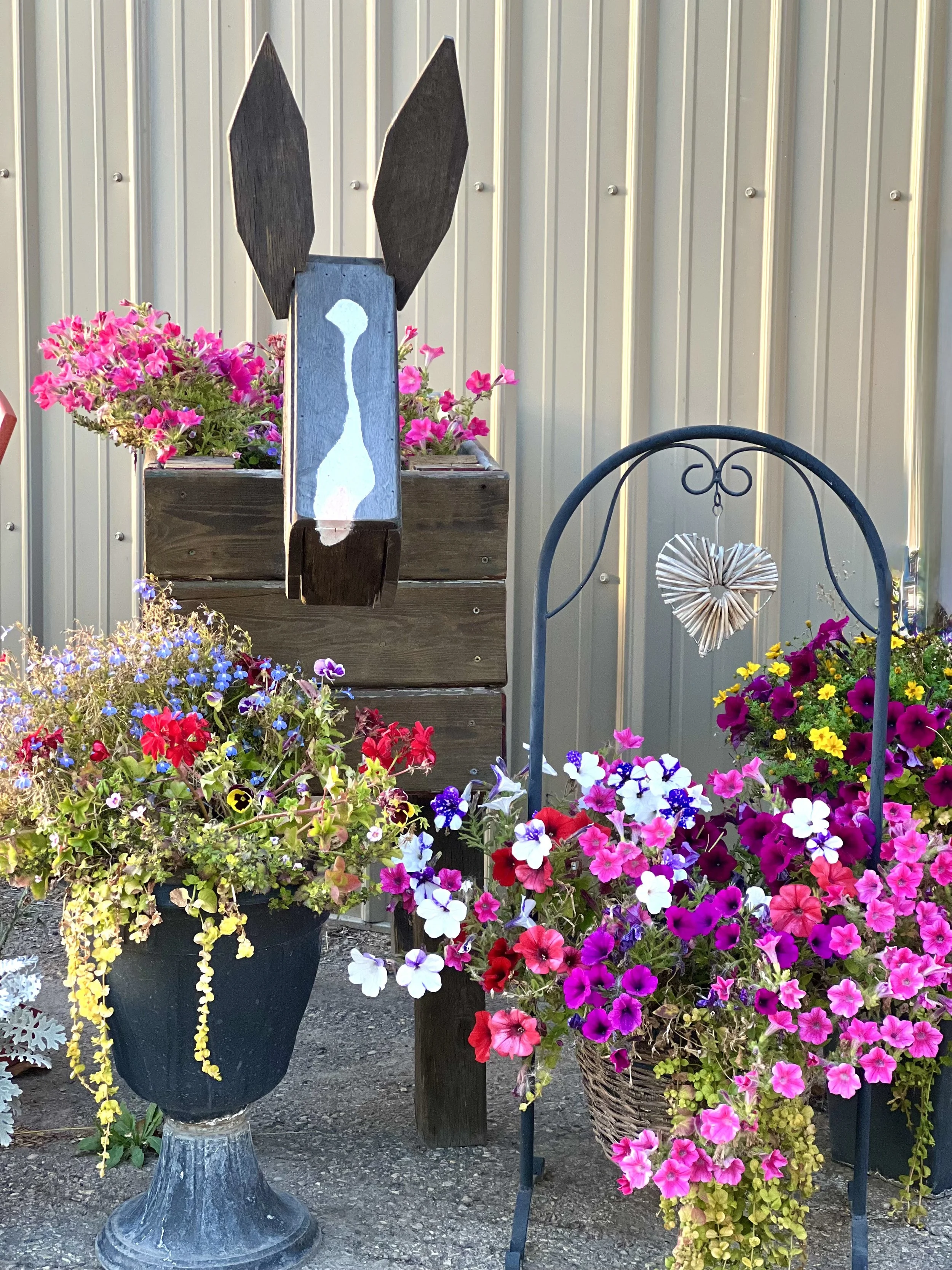 Colorful flower arrangements with pink, purple, yellow, and white blossoms, a black metal heart-shaped hanging decoration, and a wooden bunny cutout with painted white face, all set against a corrugated metal background.