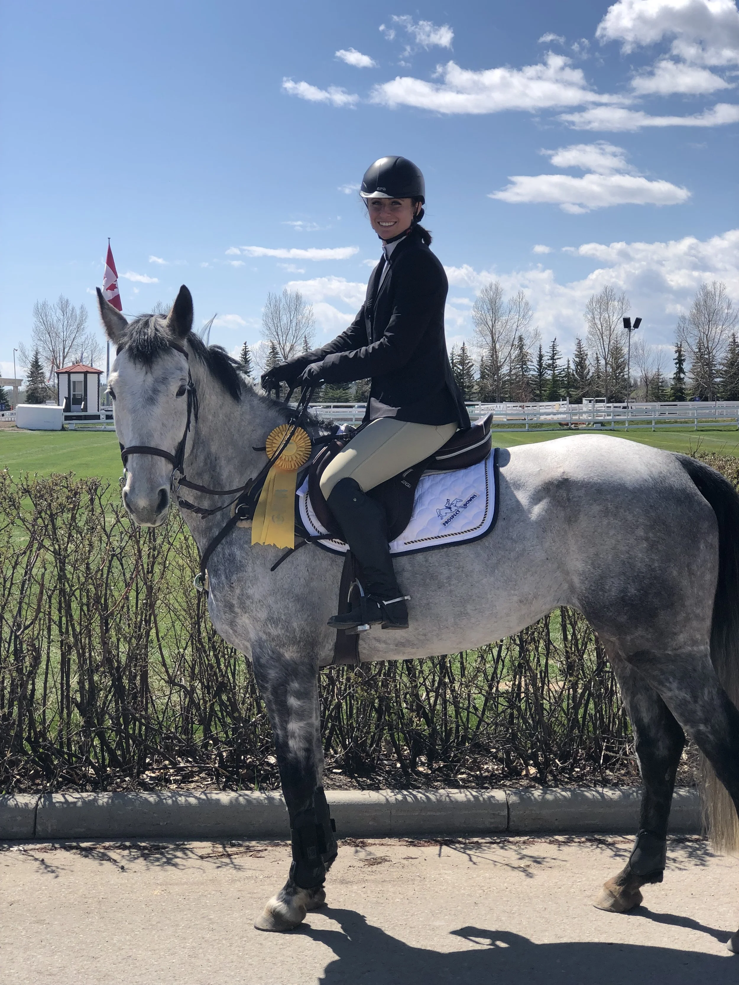A woman in equestrian attire riding a gray horse on a paved path in a park-like setting with a clear blue sky, some clouds, and a Canadian flag in the background.