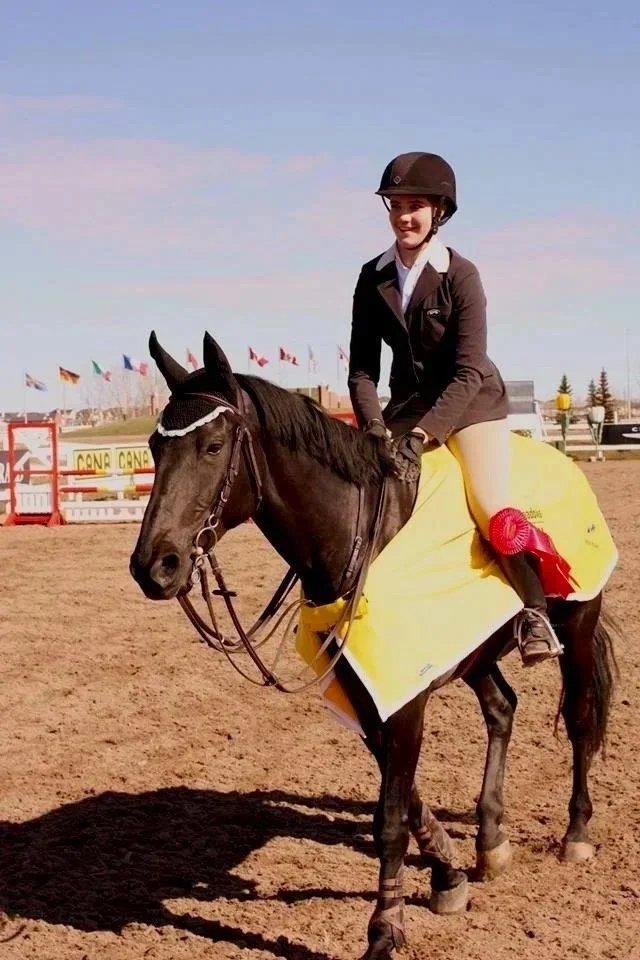 A young female equestrian rider sitting on a black horse, smiling, wearing a helmet and riding attire, at an outdoor competition with flags in the background.