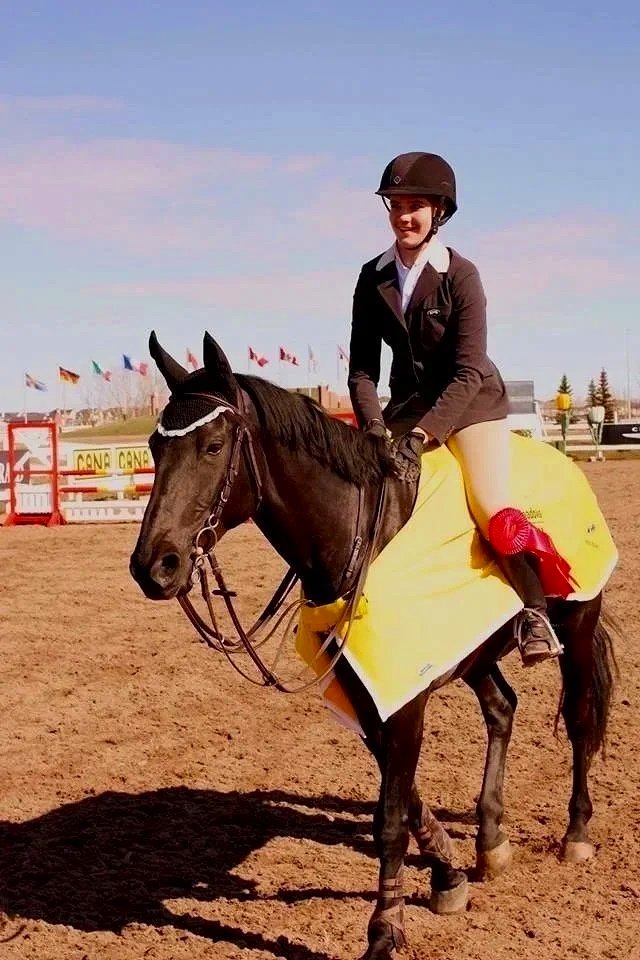 A young woman in equestrian gear sits on a brown horse with a yellow saddle blanket and a red ribbon, inside a horse riding arena.