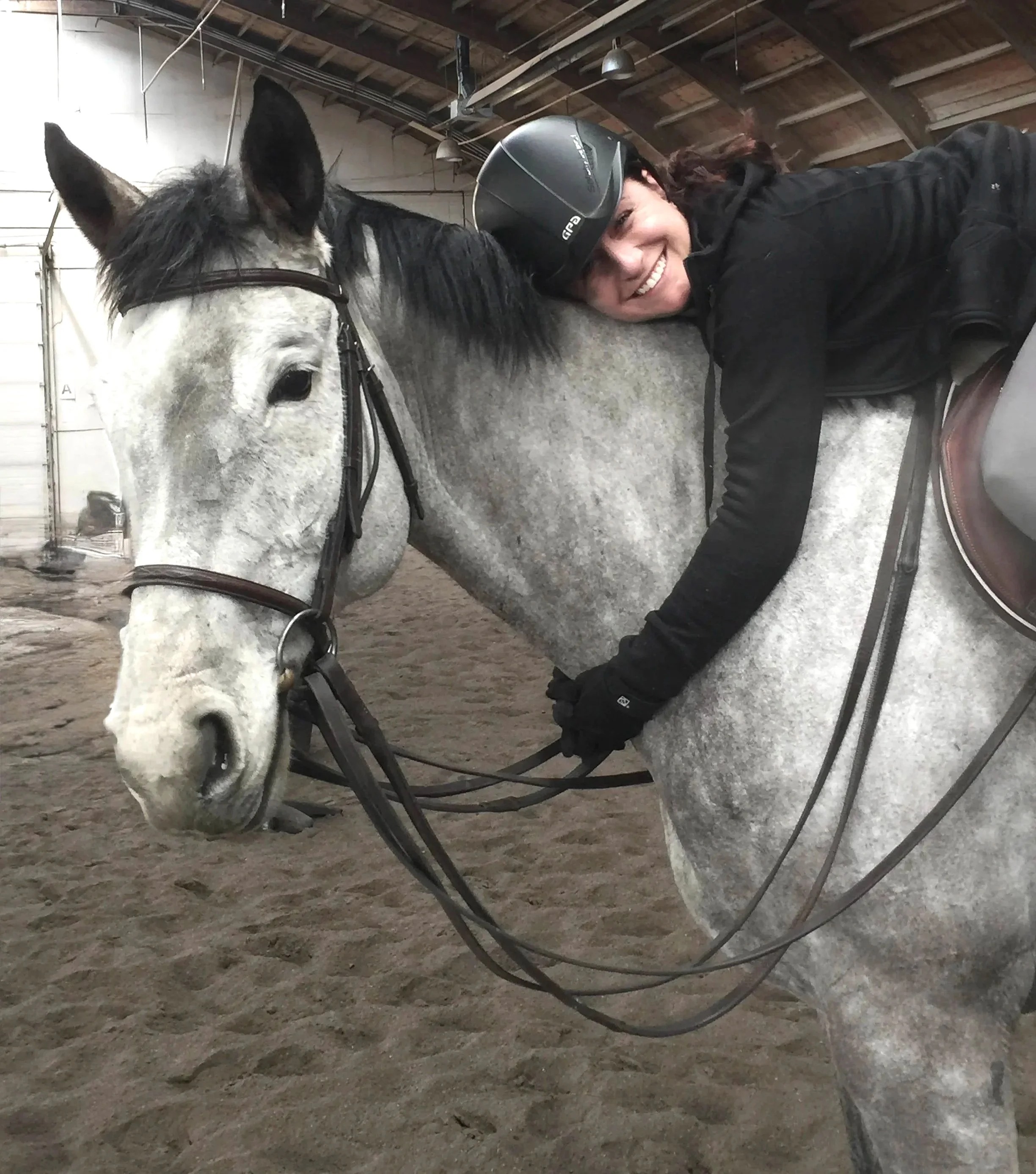 A woman with a helmet smiling and hugging a white horse inside a barn.