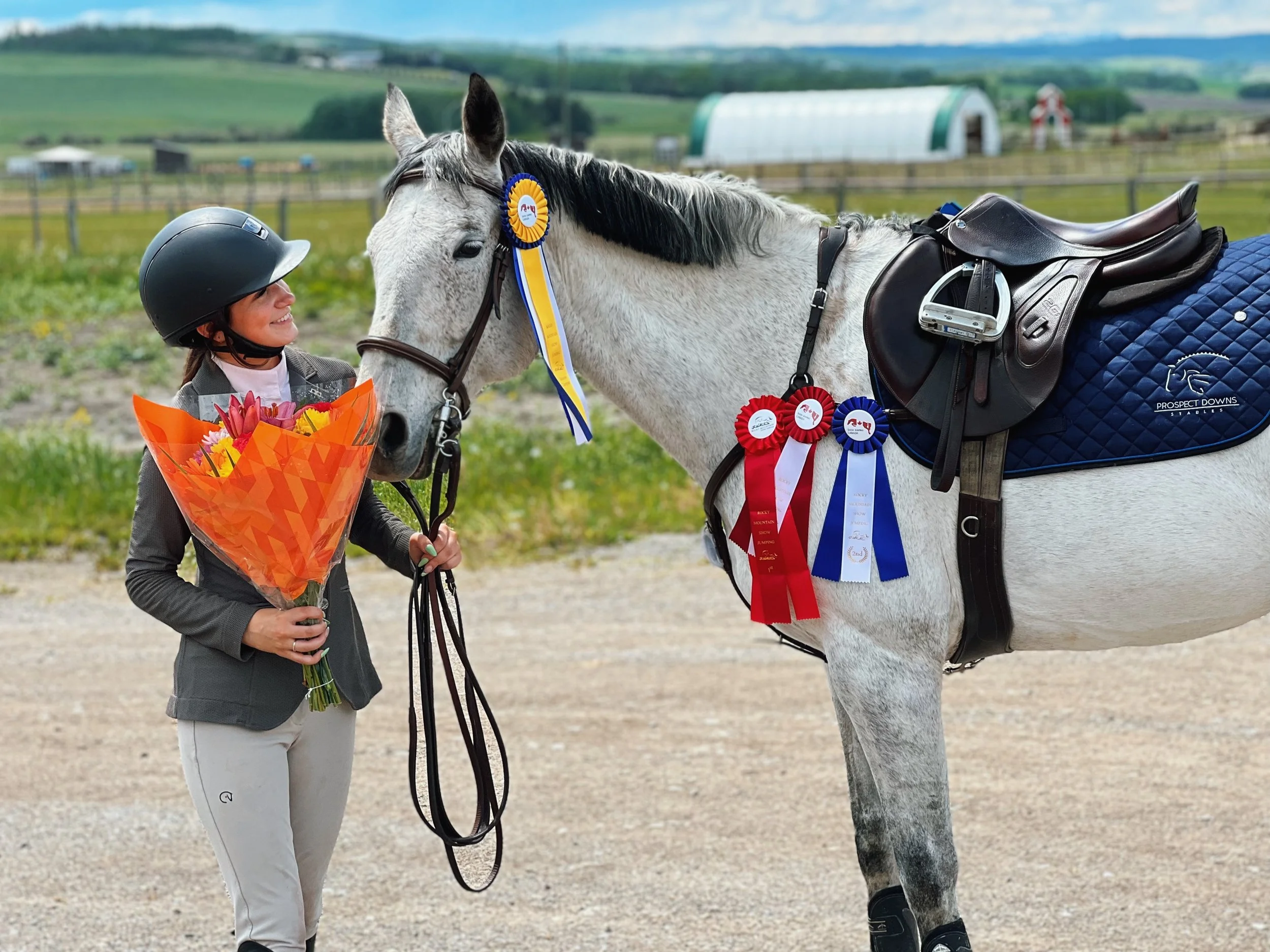 A woman in a riding helmet smiles holding a bouquet of colorful flowers while standing next to a white horse in a pasture. The horse has blue, red, and white ribbons on its bridle and a saddle. The background shows green hills, a farm building, and a