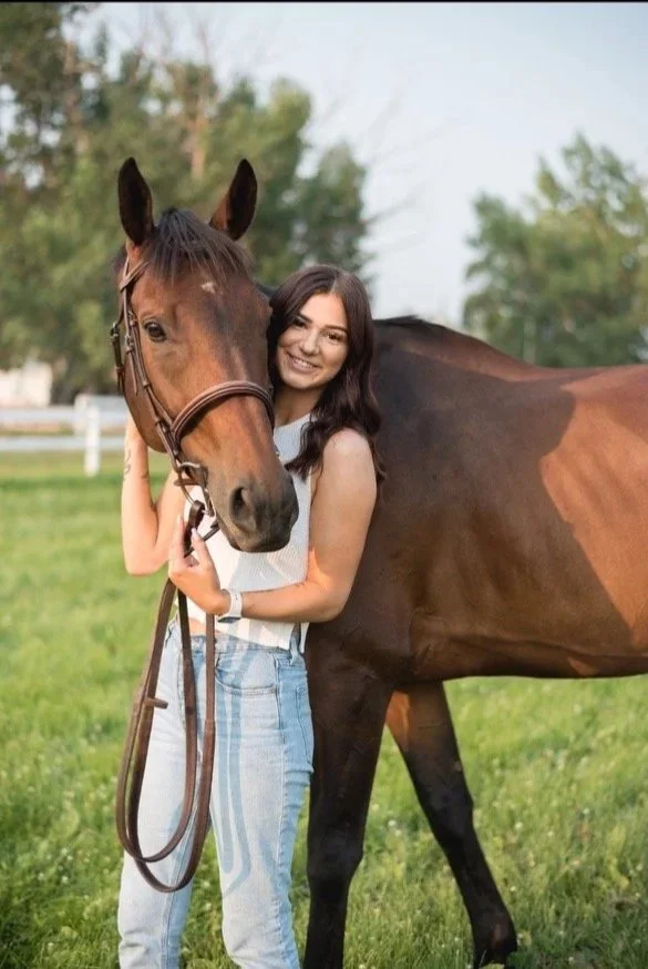 A young woman with long dark hair smiling, standing next to a brown horse on a grassy field with trees in the background.