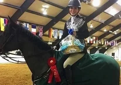 A woman wearing a helmet and riding a horse inside an indoor equestrian arena. She is holding a large gift bag and is seated on a horse adorned with a red ribbon, indicating a victory or award.