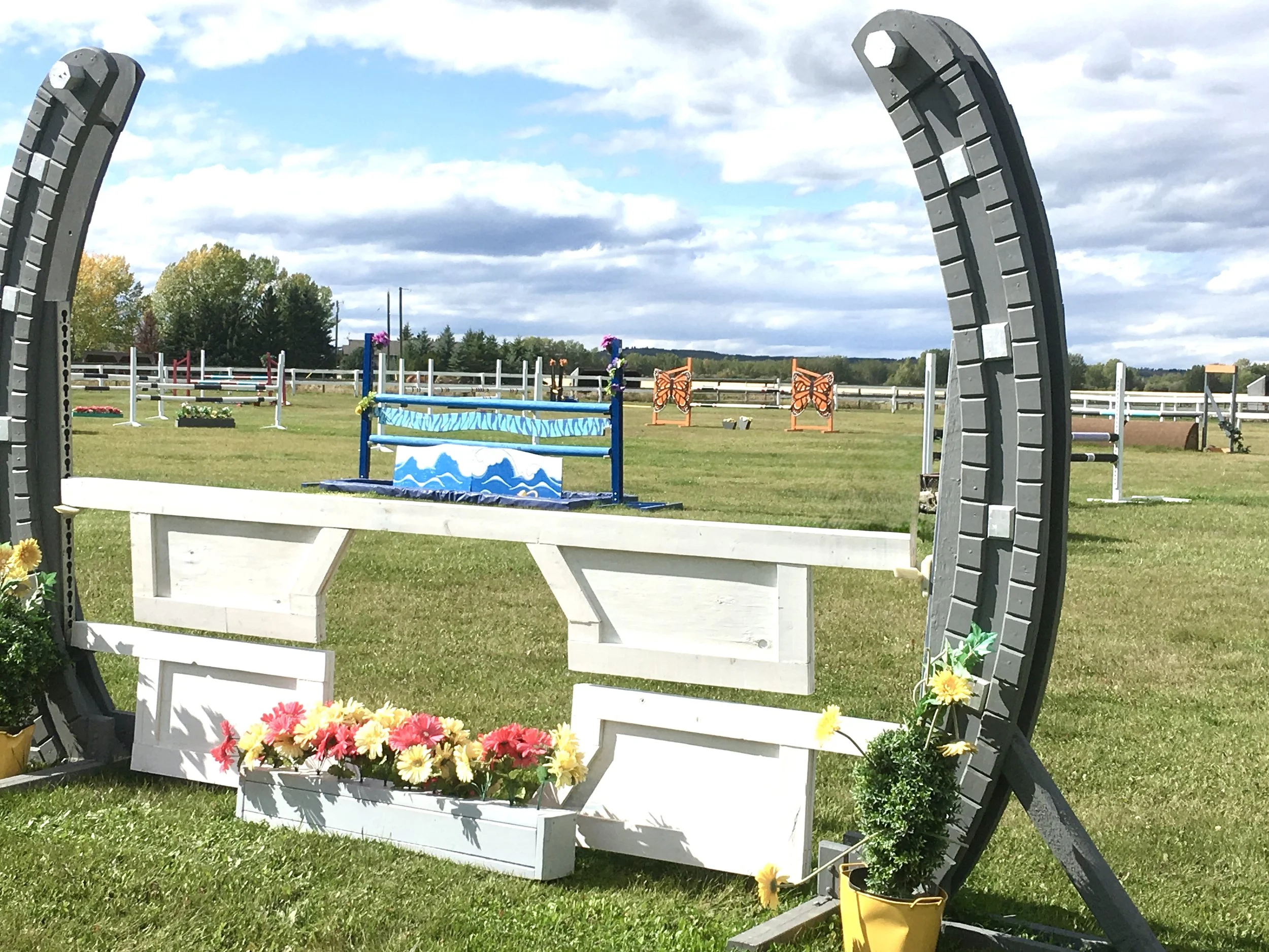 An outdoor horse jumping arena view from a decorative black circular frame with a white wooden bench in front, a flower bed, and colorful jumps set up on a grassy field with trees and a cloudy sky in the background.