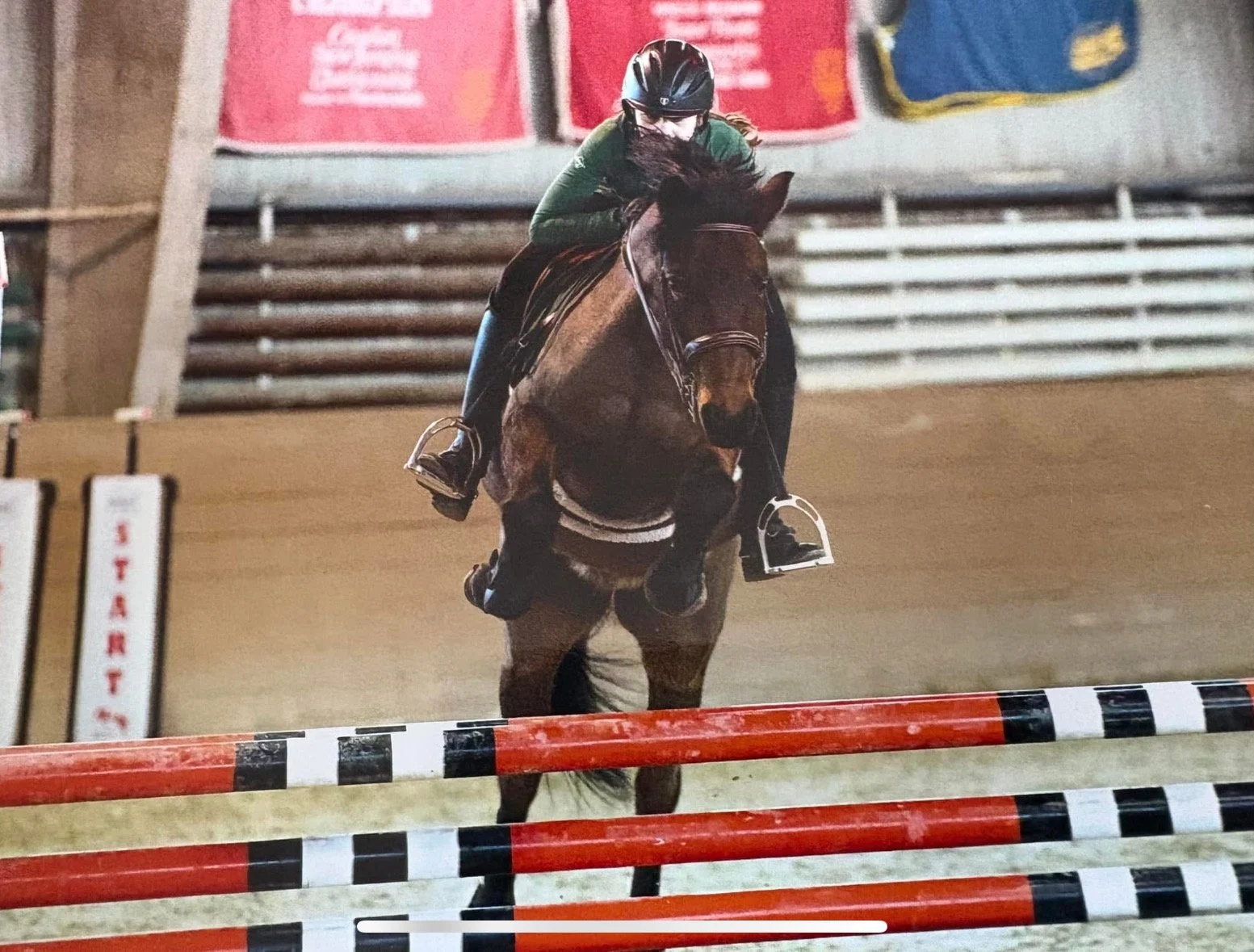 A female jockey riding a brown horse during a gymkhana event, jumping over a red and white striped obstacle on an indoor dirt track.