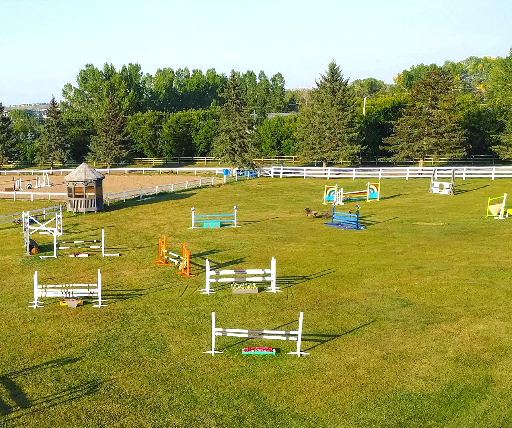 An outdoor space with various colorful equestrian jump obstacles on a well-kept green grass field, surrounded by a white fence and tall trees in the background.