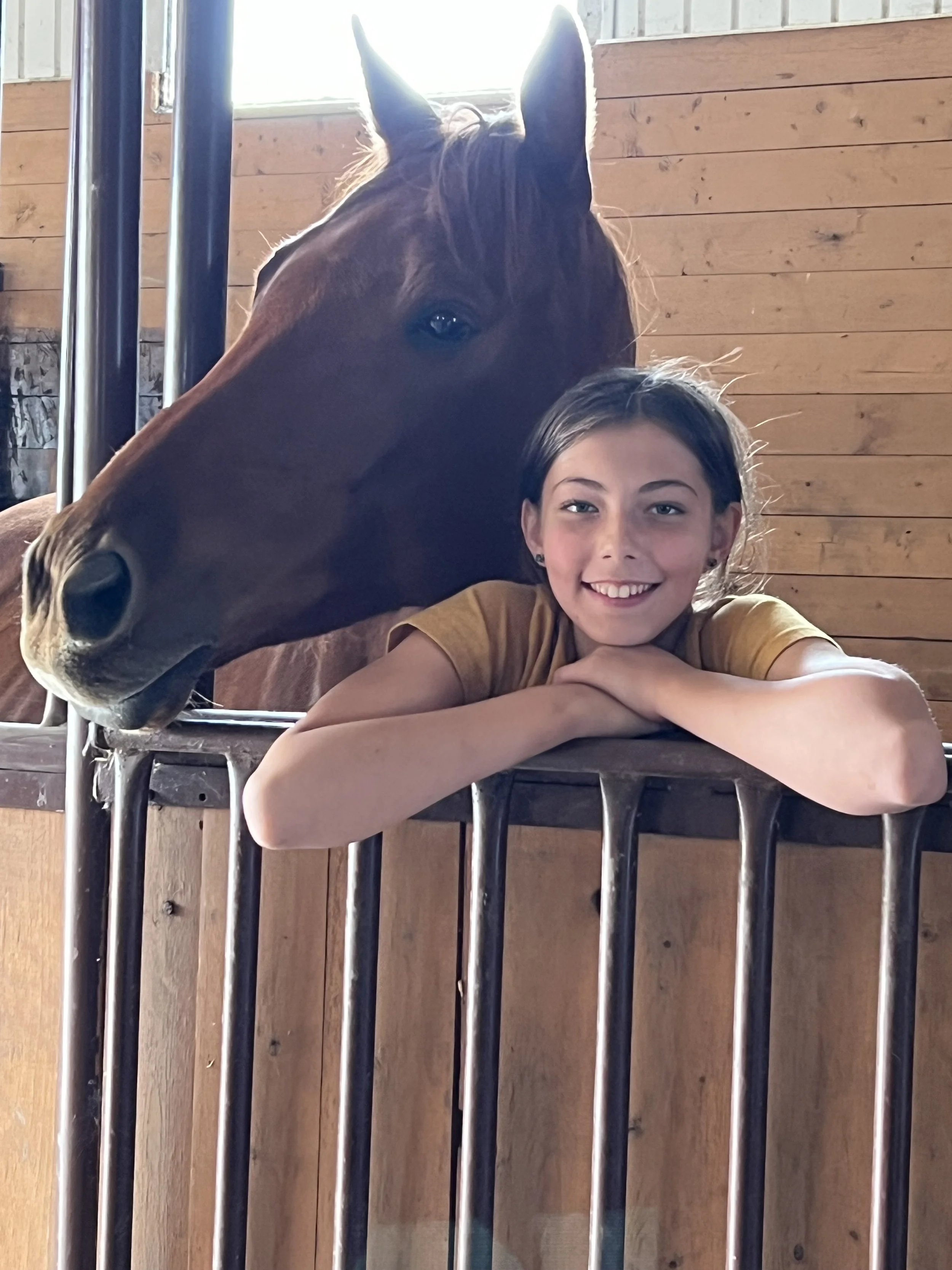 A girl with a big smile resting her chin on her crossed arms, leaning on a wooden and metal railing, with a large horse's head close to her, inside a stable with wooden paneling.