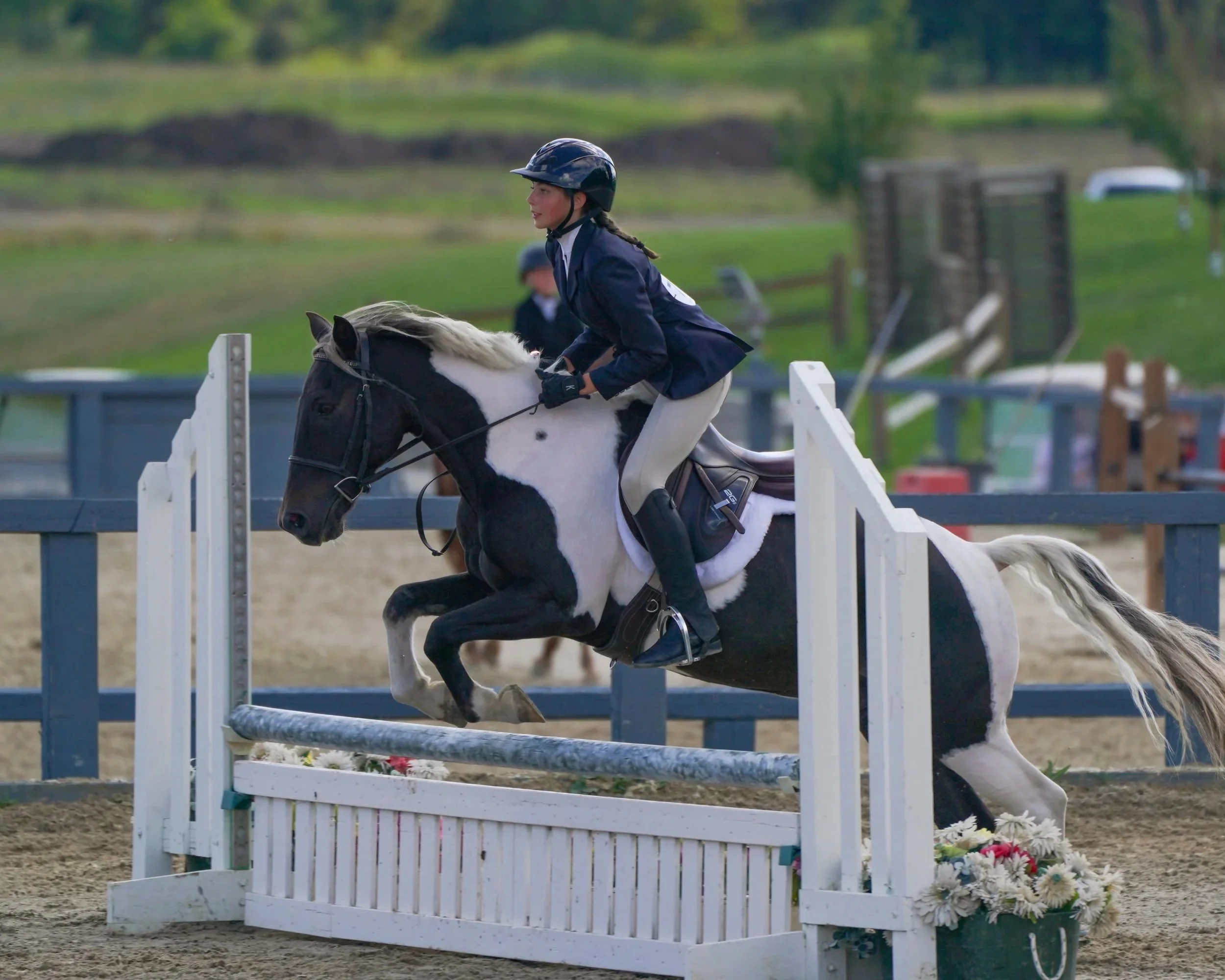 A female equestrian rider wearing a helmet and formal riding attire guides her horse over a jumping obstacle at an outdoor equestrian event, with green fields and training facilities in the background.
