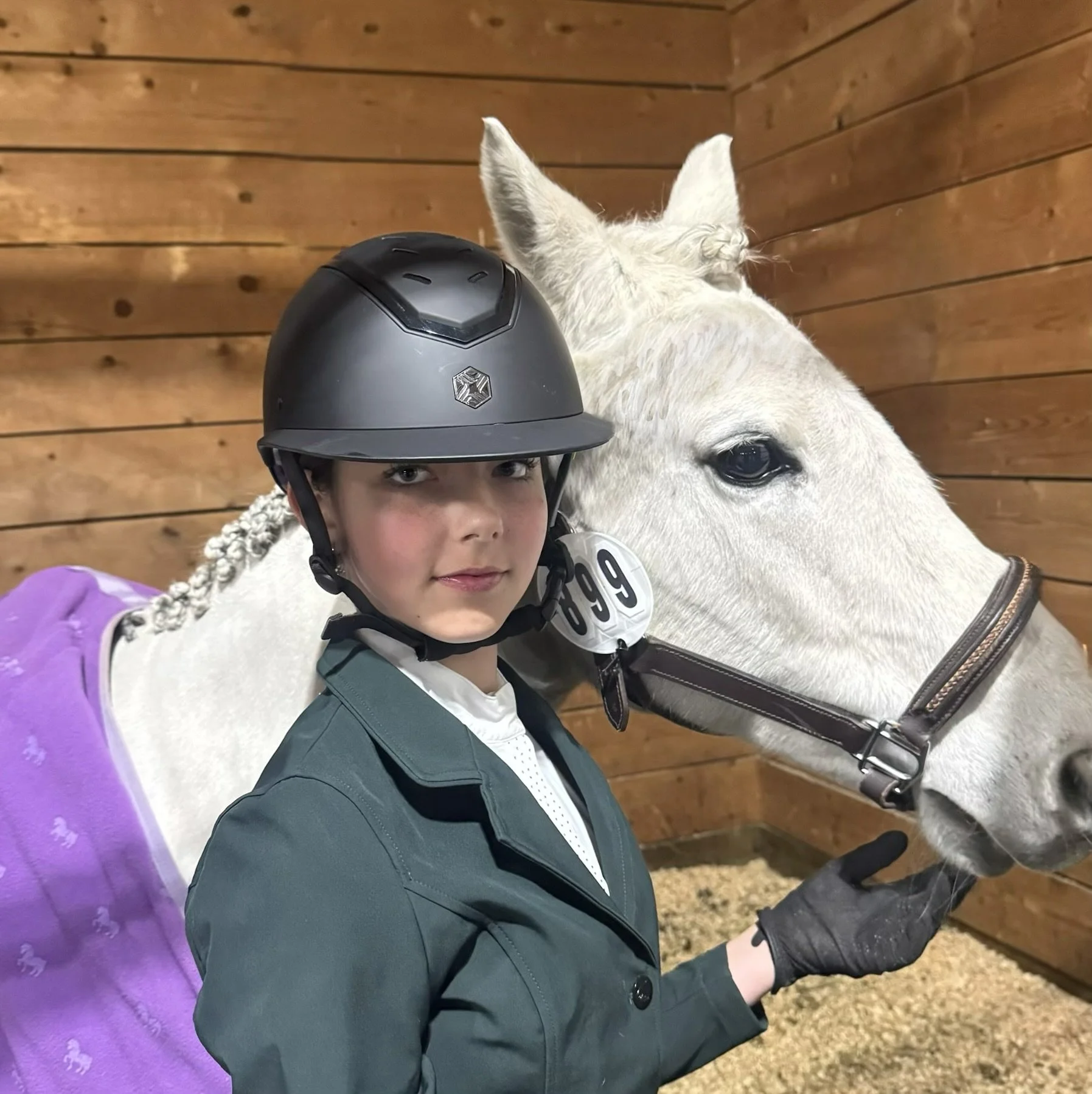 A person wearing a riding helmet and riding jacket stands next to a white horse inside a wooden indoor stable.