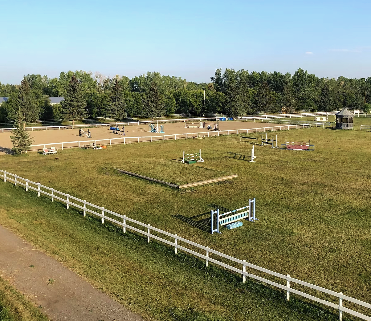 An outdoor equestrian training or jumping arena with various horse jumps and structures, surrounded by white fencing and green trees in the background.
