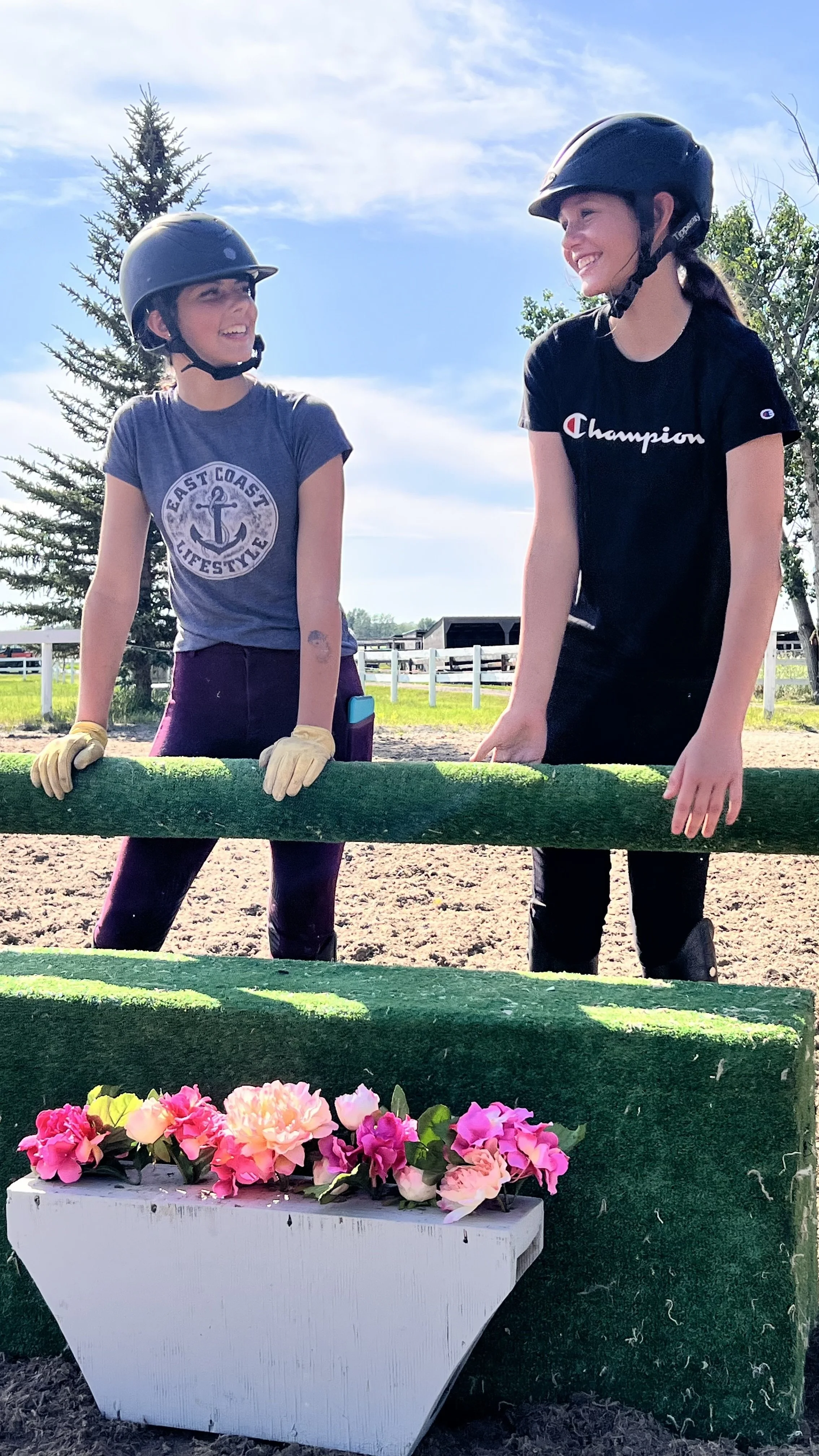 Two women wearing helmets and gloves, standing outdoors near a fence, smiling and talking to each other, with pink flowers in a white planter in front of them.