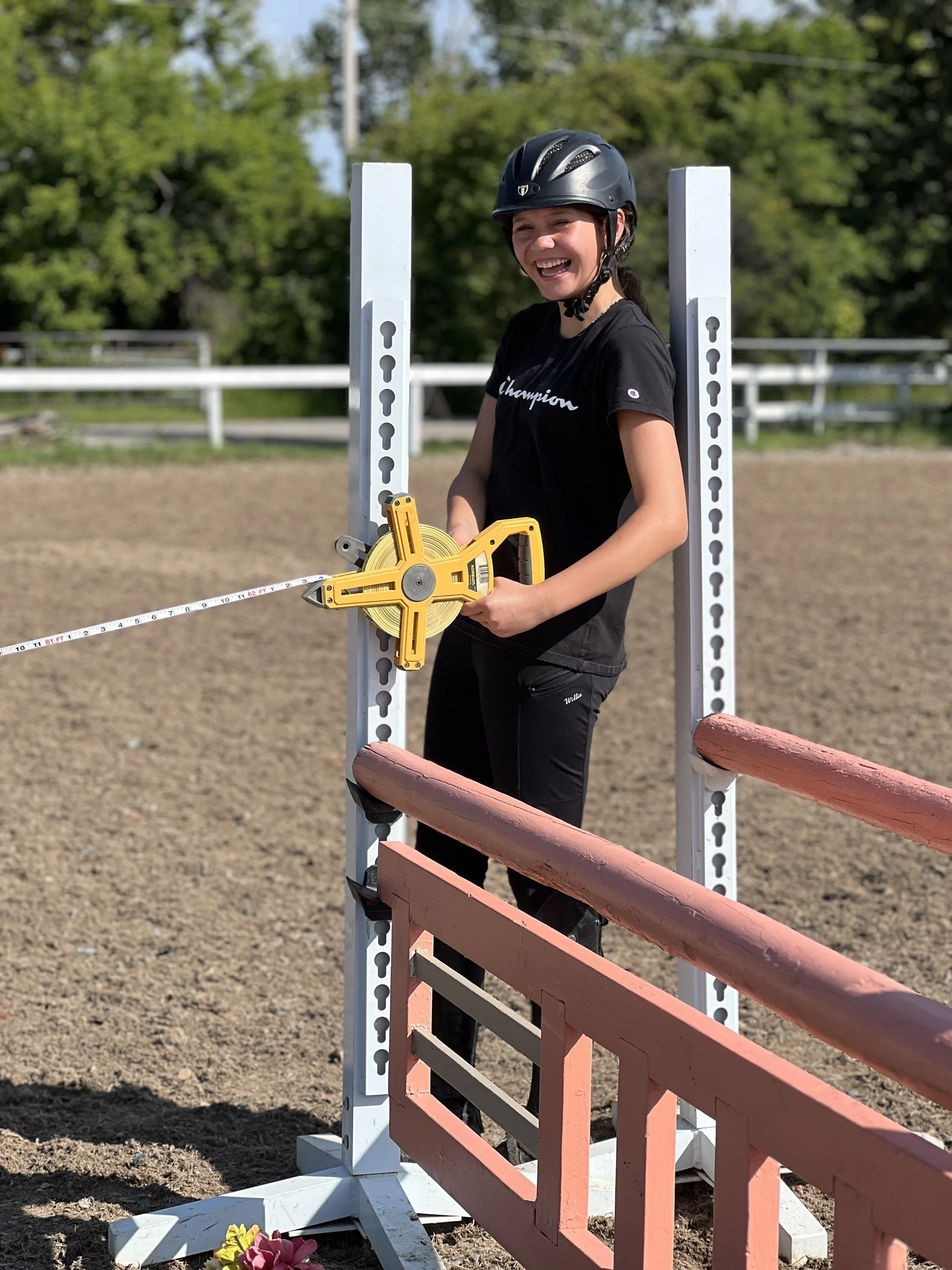 A young girl with a helmet smiling and holding a tape measure at an outdoor horse jumping arena, with a pink and train of flowers at the base of the jump.