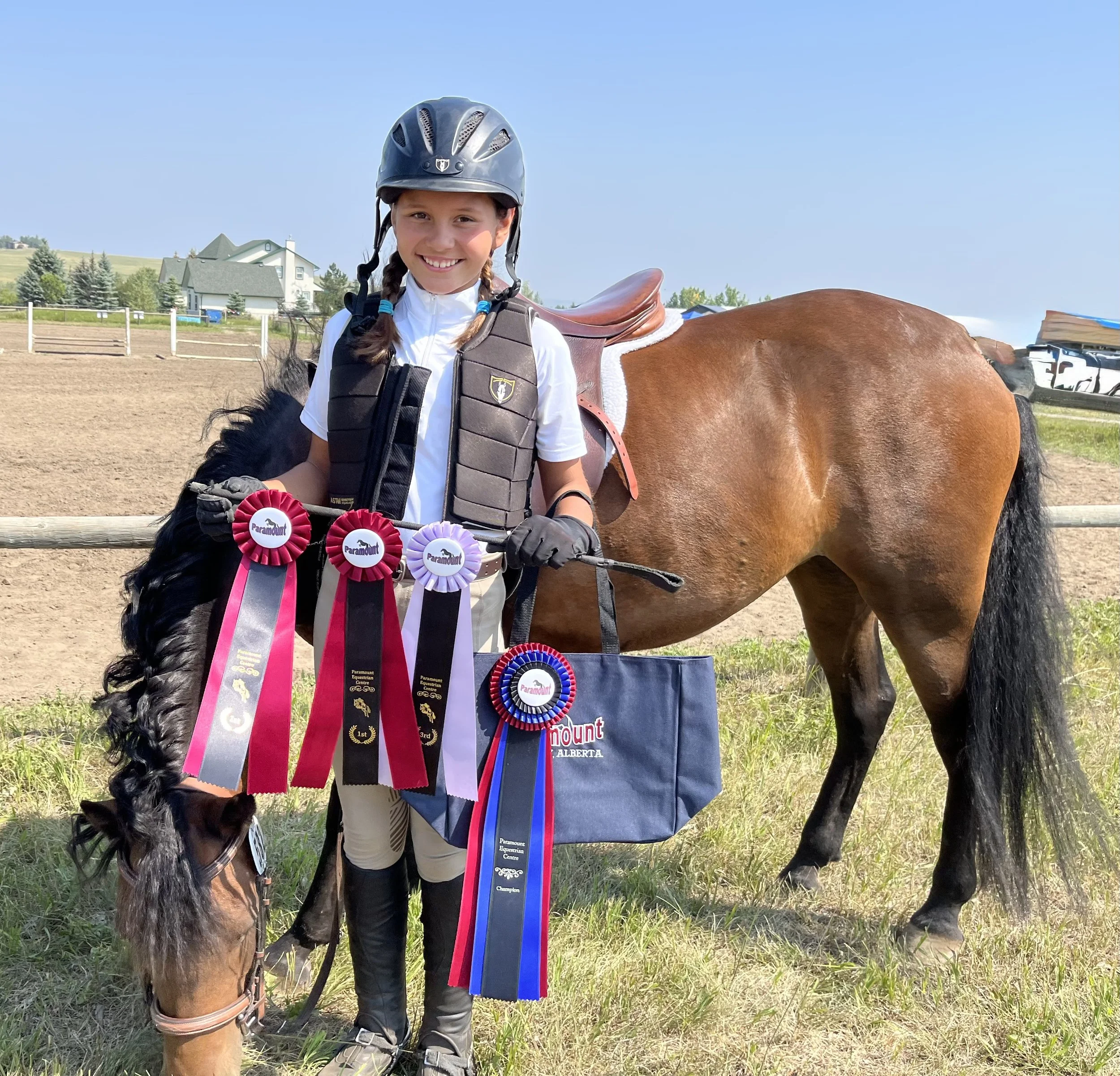 A young girl smiling in equestrian attire, including a helmet and protective vest, standing beside a brown horse with ribbons attached, at an outdoor riding arena on a sunny day, with houses and trees in the background.
