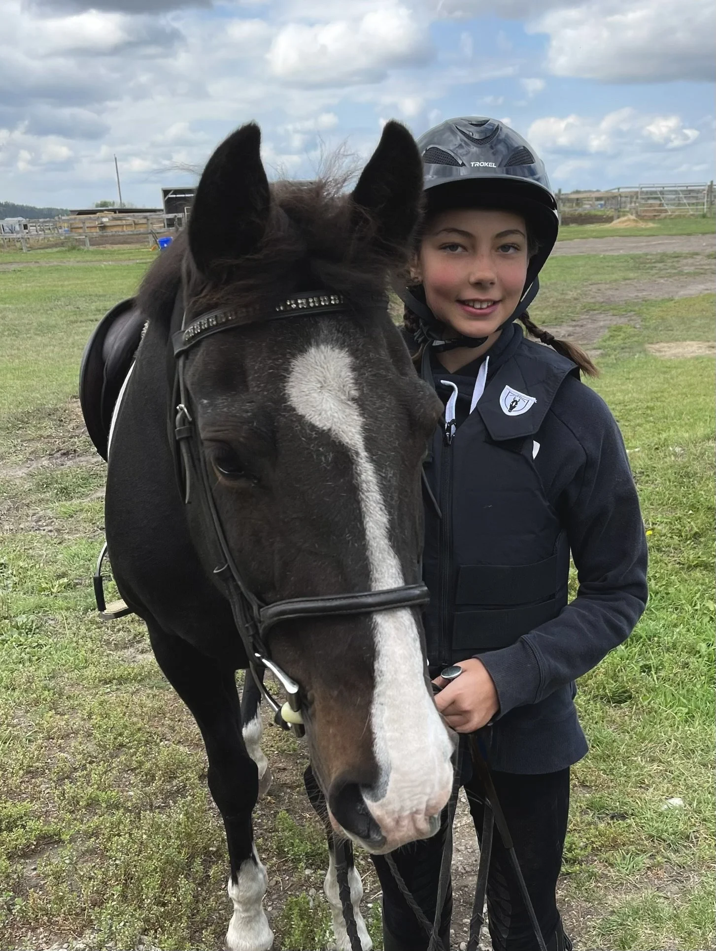 A young girl wearing a helmet and riding vest stands next to a black and white horse in an open grassy area.