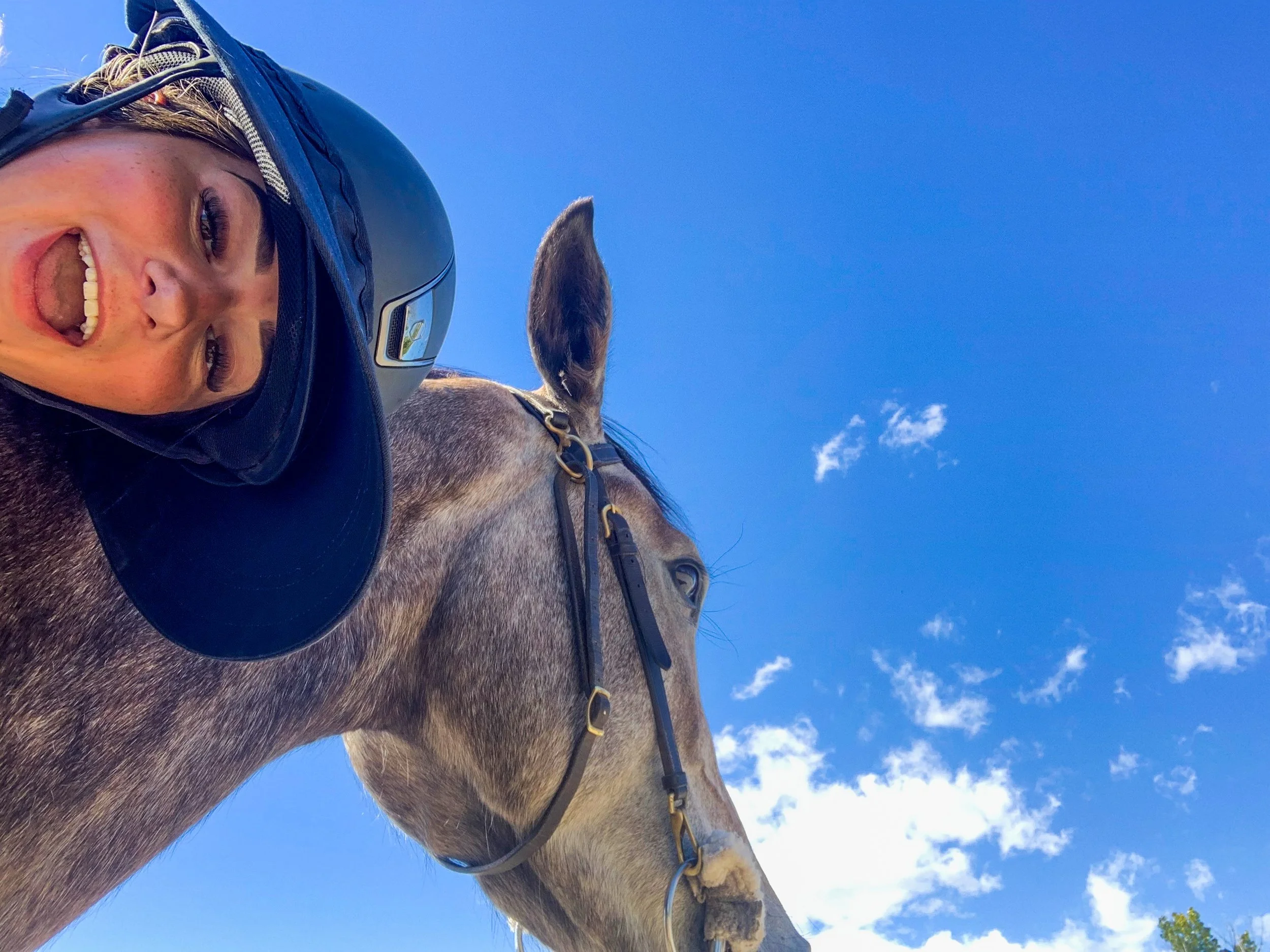 A person with long hair, wearing a riding helmet, smiling, riding a horse in a clear blue sky.