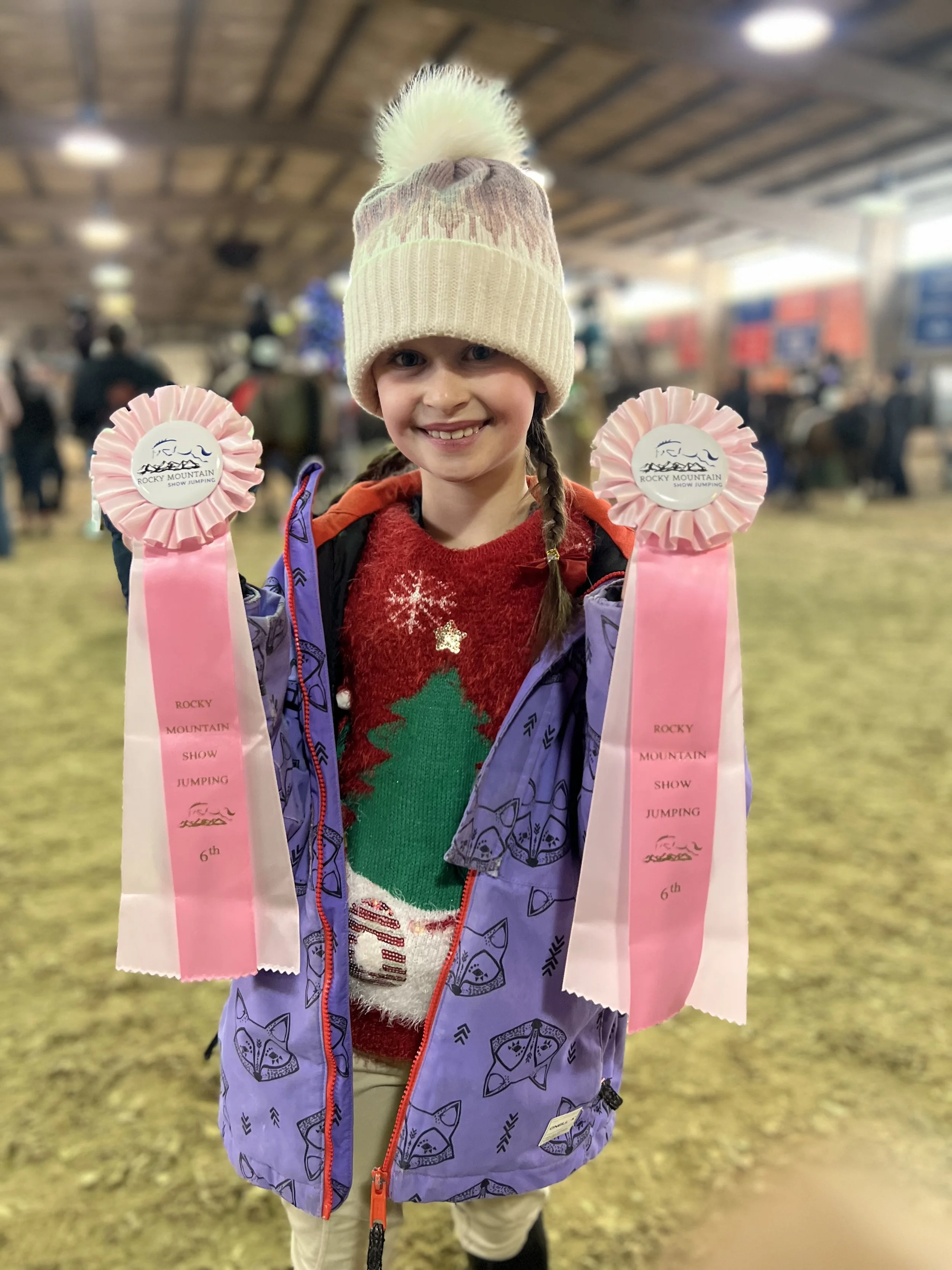 A young girl wearing a beige knit hat with a pom-pom and a purple jacket, holding pink ribbons for a show jumping competition at Rocky Mountain Show Jumping, with a smile on her face and a blurred indoor event background.