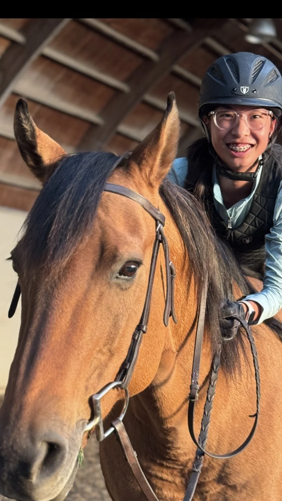 A person wearing a helmet and glasses riding a brown horse inside a barn with a wooden ceiling.
