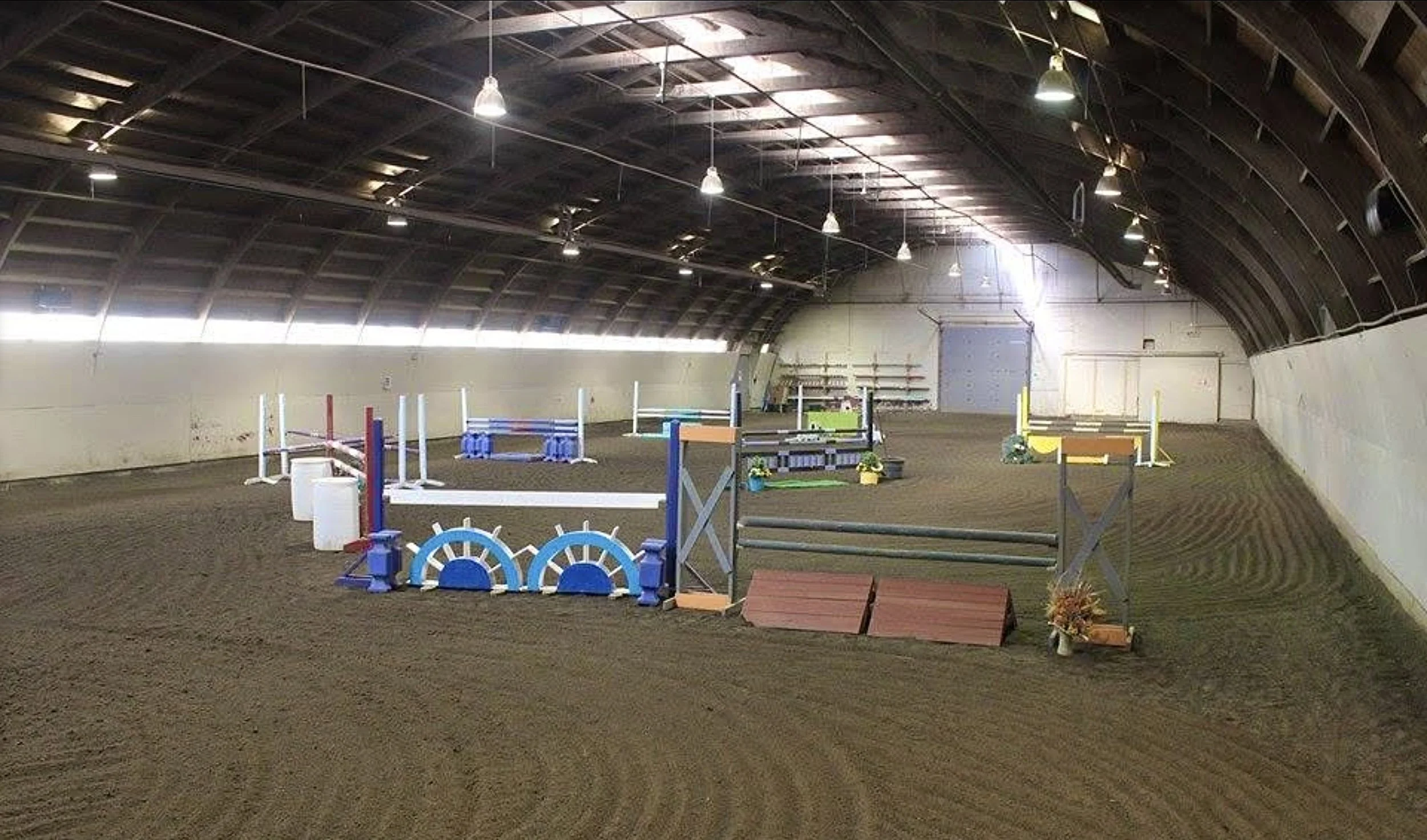 Indoor horse riding arena with jumps, poles, and decorative flowers, under a curved wooden ceiling with hanging lights.