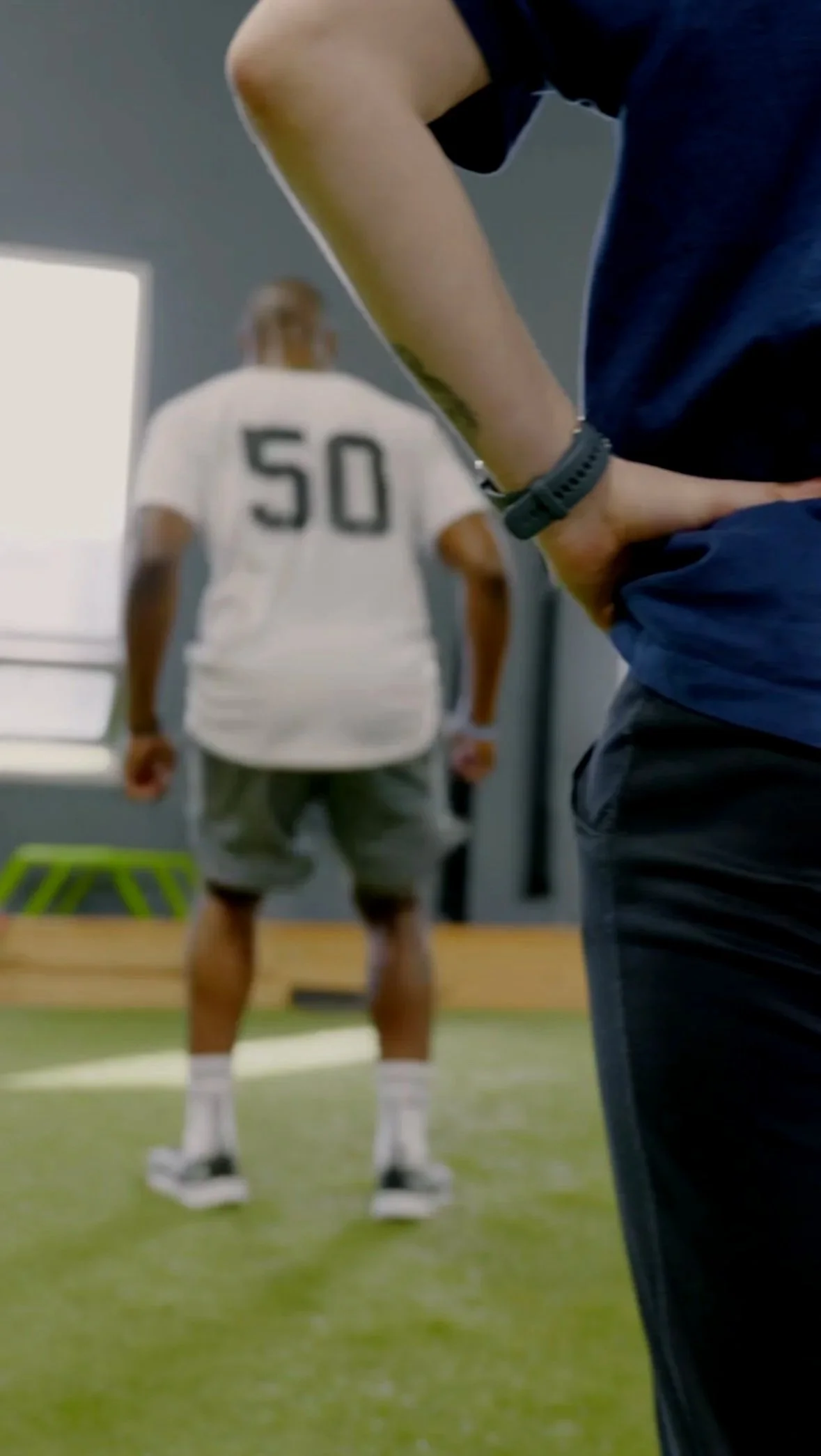 A person wearing a navy blue shirt stands on a green indoor turf, with their hand on their hip, observing a football player in a white jersey with the number 50, who is standing in the background.