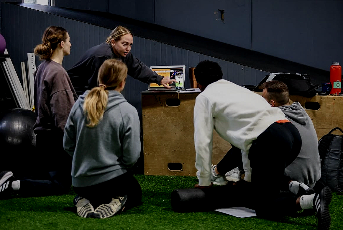 A group of five women and men participating in a fitness class or training session indoors, with one woman leading instructions on a smartphone, while others are kneeling or leaning on foam rollers on artificial grass.