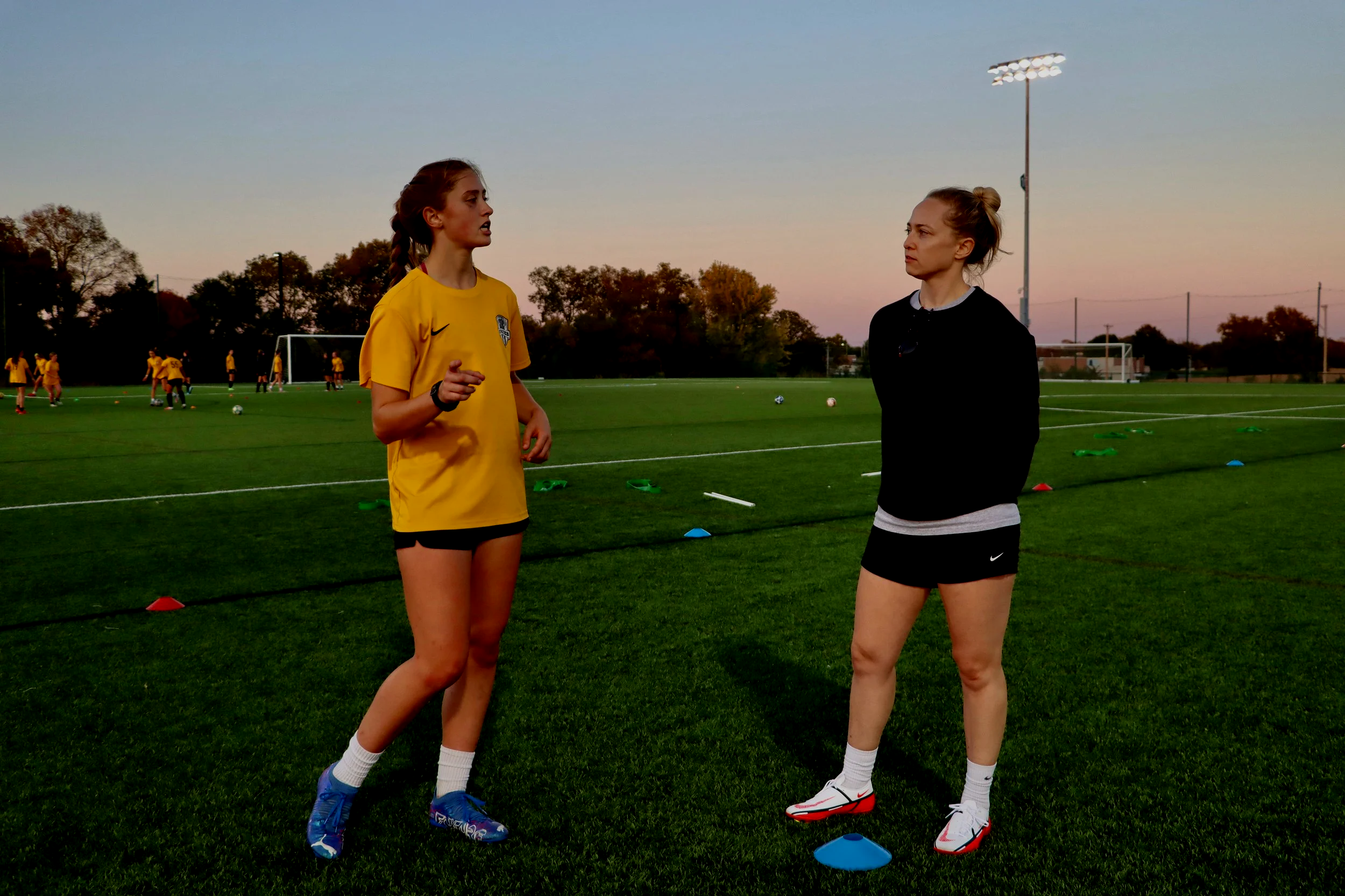 Two female soccer players standing on a soccer field during practice, one wearing a yellow jersey and the other dressed in black, with multiple players in yellow jerseys practicing in the background at sunset.