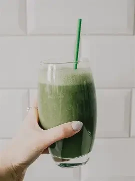 A hand holding a glass of green smoothie with a green straw, against a white background.