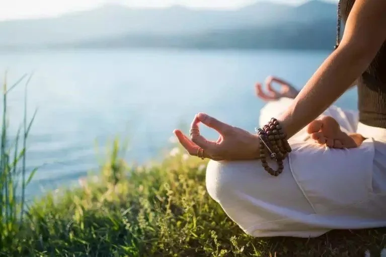 Person practicing meditation outdoors near a lake, sitting cross-legged on grass with hands in a mudra position.