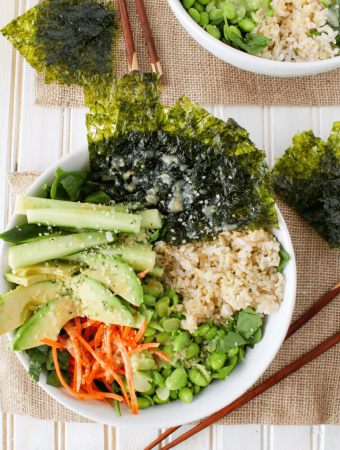 A bowl of mixed Asian-inspired salad with seaweed, cucumber, shredded carrots, edamame, rice, and sesame seeds, with a side bowl of rice and peas.