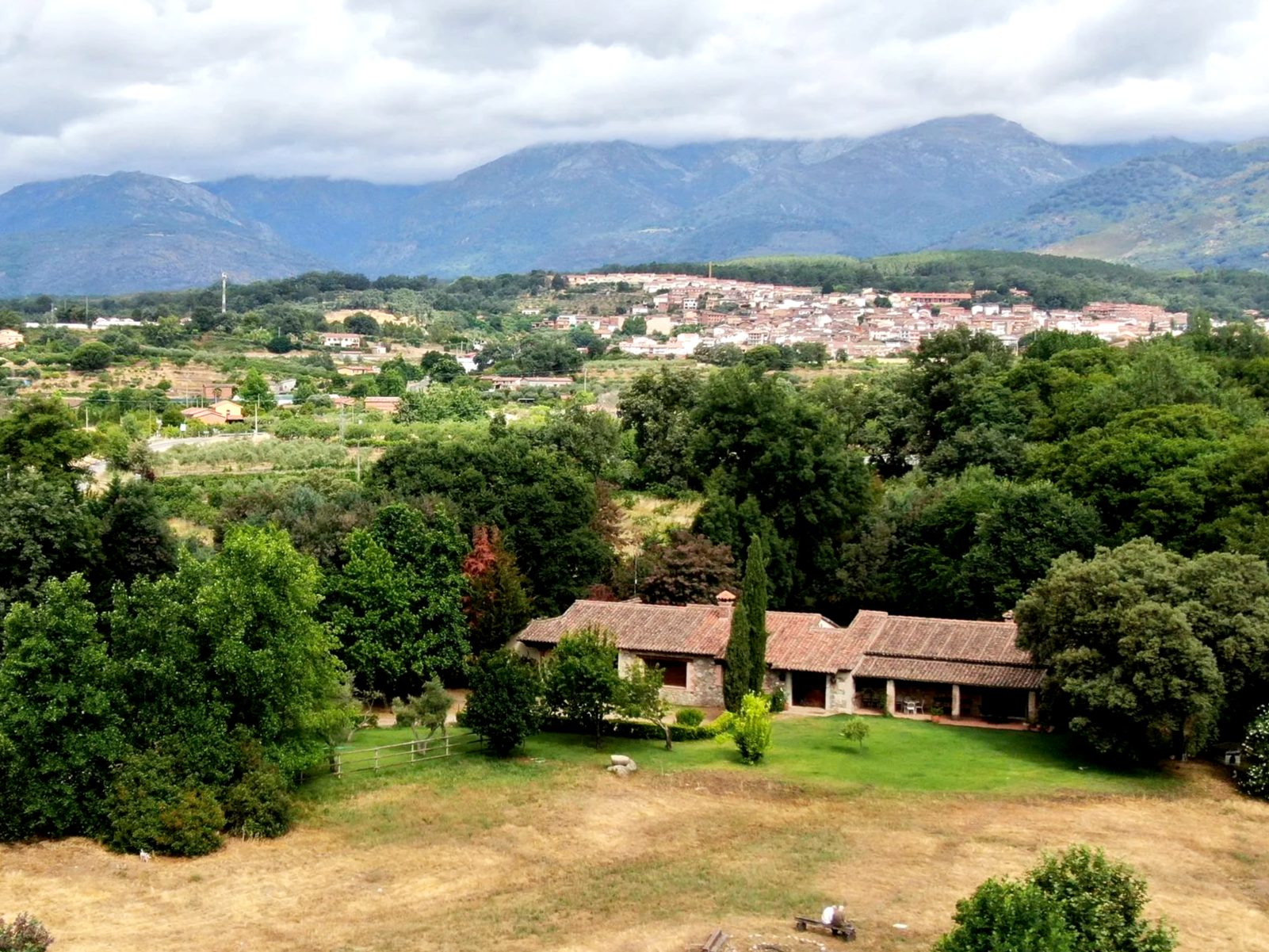 Paisaje de colinas verdes y montañas, con un pueblo en la distancia y una casa antigua rodeada de árboles y césped en primer plano.