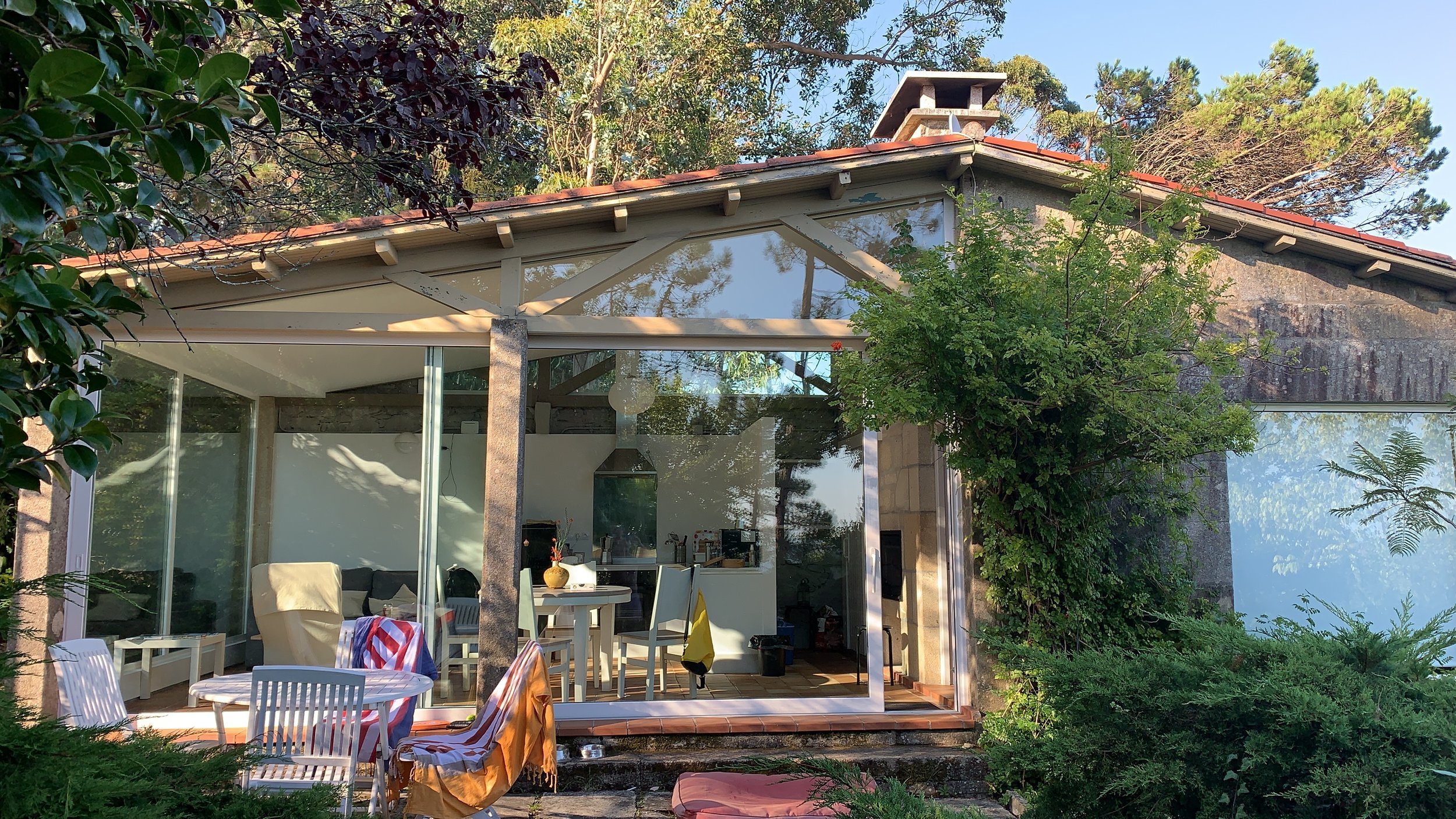 Casa moderna con paredes de cristal y techo de tejas rojas, rodeada de árboles y vegetación, con un área exterior con sillas y mesa