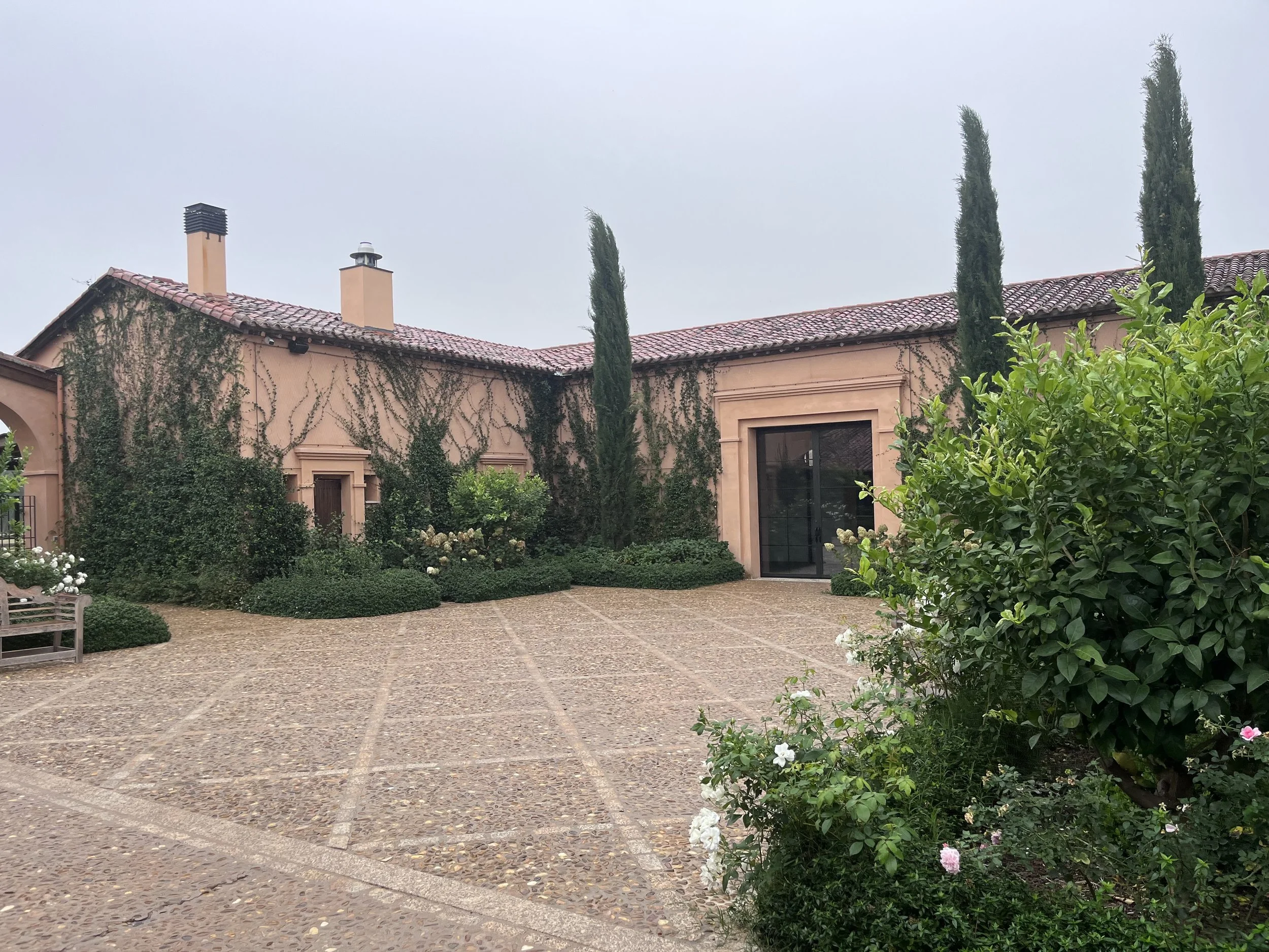 Casa con paredes de color beige, tejado de tejas rojas y ventanas con marcos negros, rodeada de plantas y árboles en un patio pavimentado con piedras.
