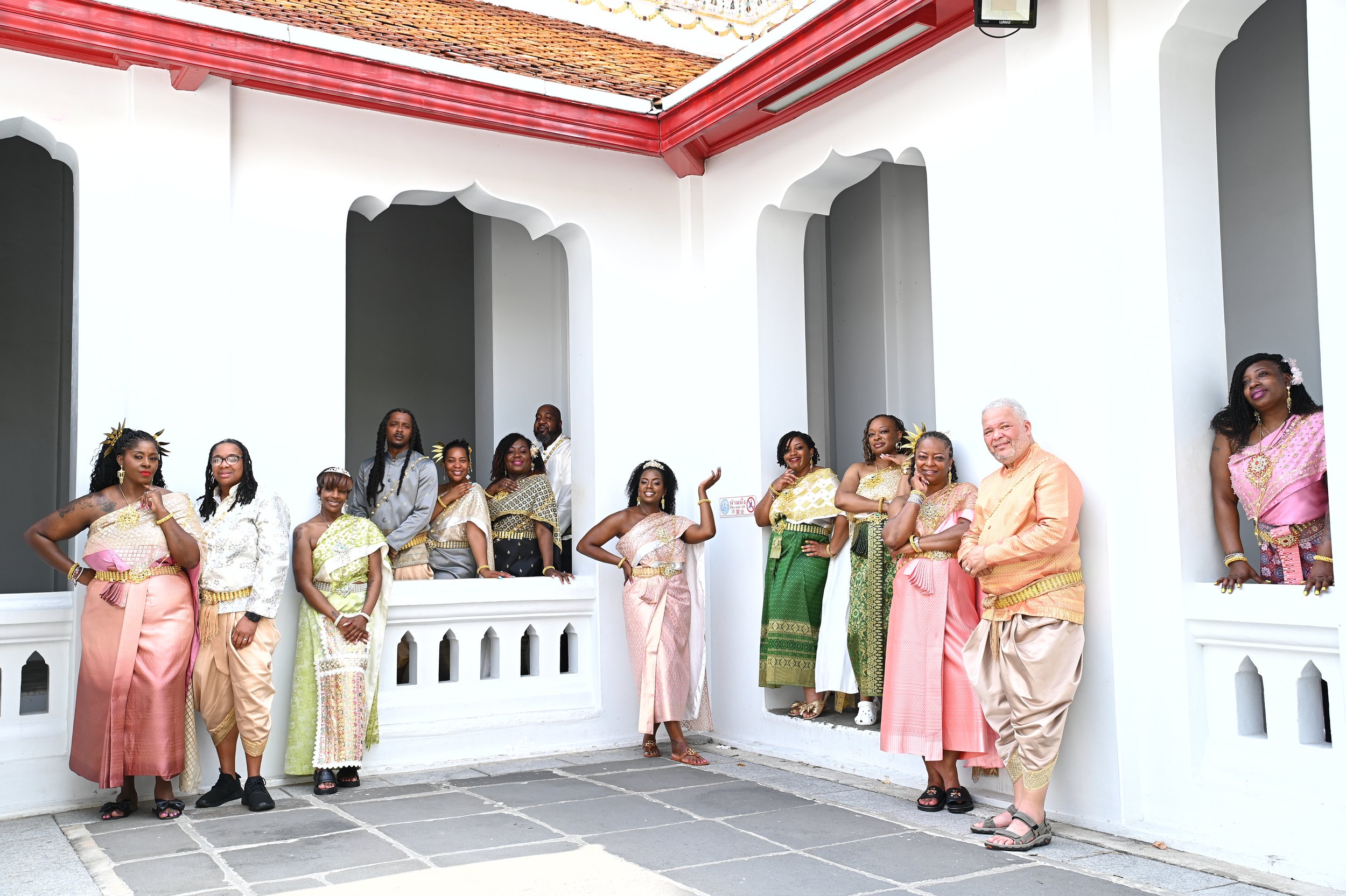 Group of people dressed in traditional and colorful clothing standing against a white wall of a building with arched openings, sunny weather, and tiled roof.