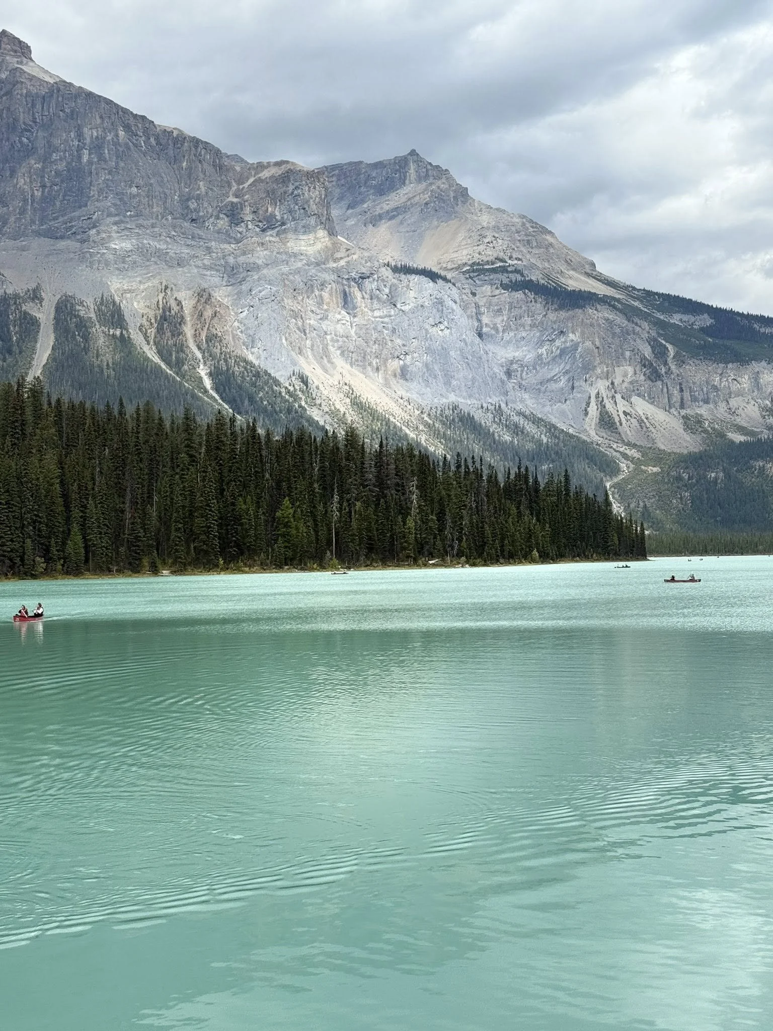 Scenic landscape of a turquoise lake surrounded by pine trees and mountainous cliffs, with a cloudy sky overhead and a few small boats on the water.