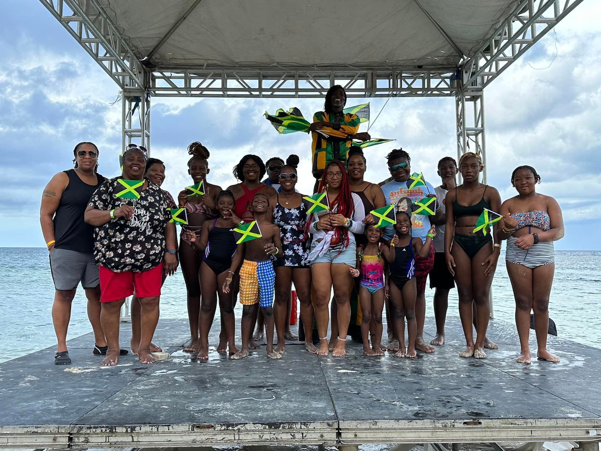 Group of people on a stage at the beach, holding Jamaican flags, smiling, with some children, a person standing on top of the stage, and ocean in the background.