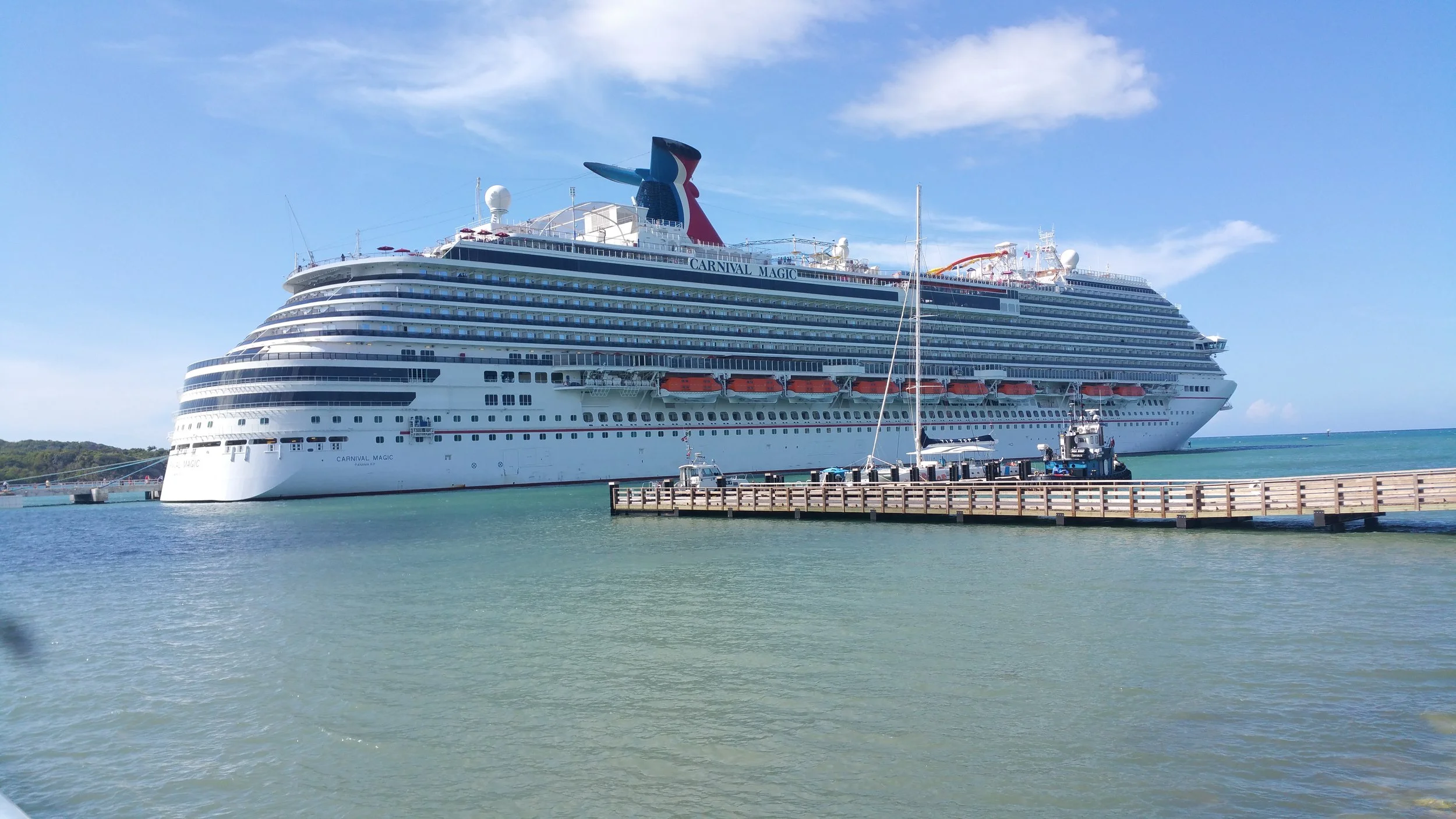Large white cruise ship named Carnival Magic docked at a pier in calm turquoise water under a partly cloudy sky.