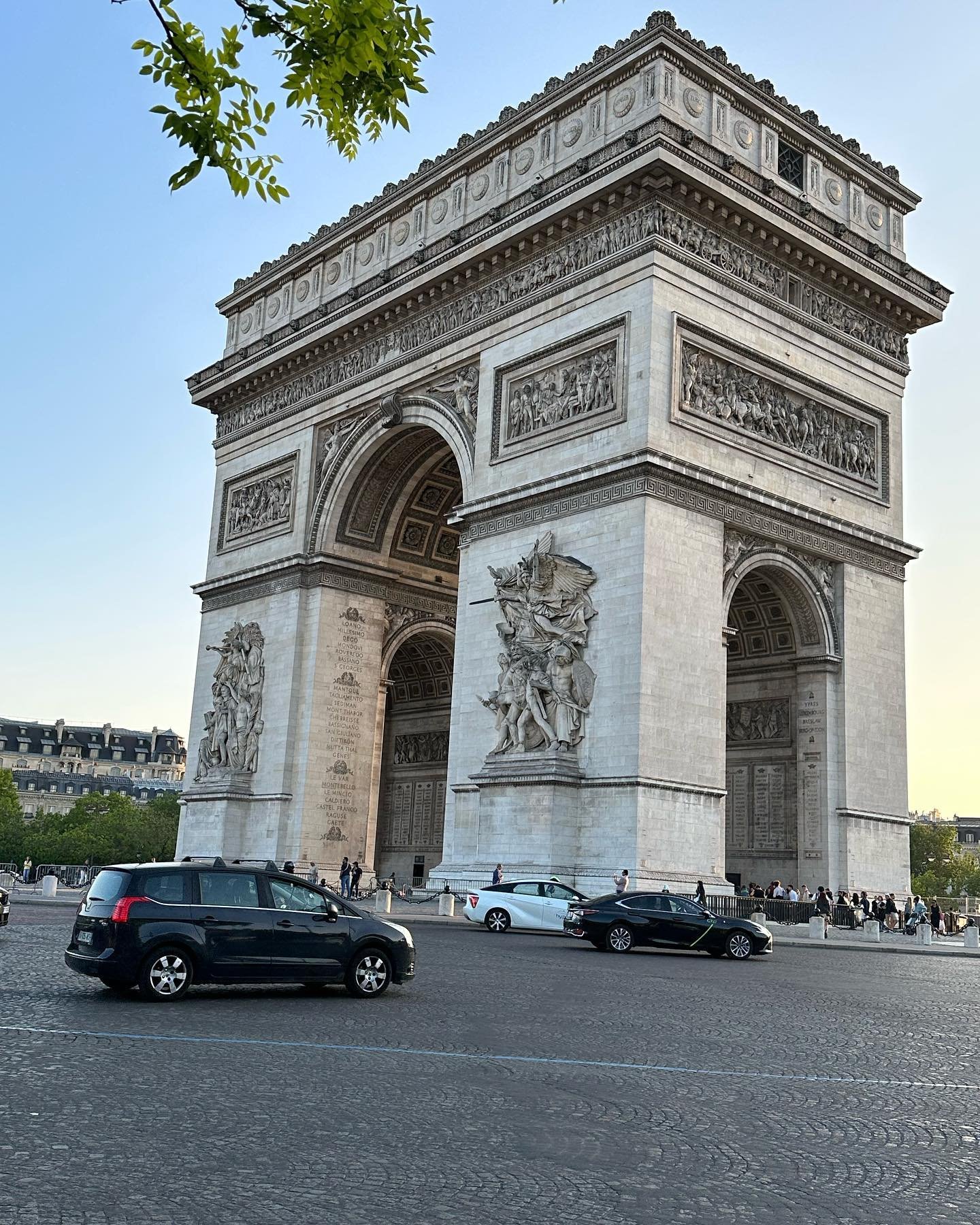 The Arc de Triomphe, a large historic monument in Paris, France, with cars driving on the road in front and people walking around the base, under a clear sky.
