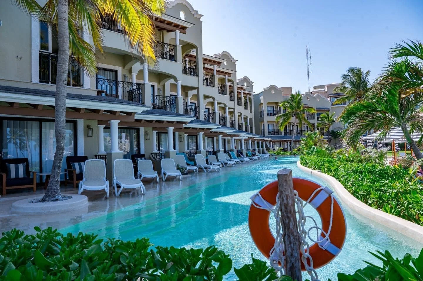 Pool area at a tropical resort with lounge chairs, palm trees, and a hotel building in the background.