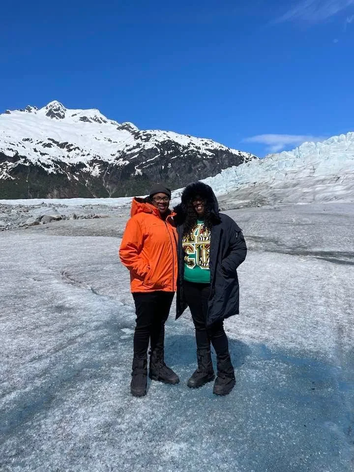 Two women in winter jackets standing on ice with snow-capped mountains and glaciers in the background.