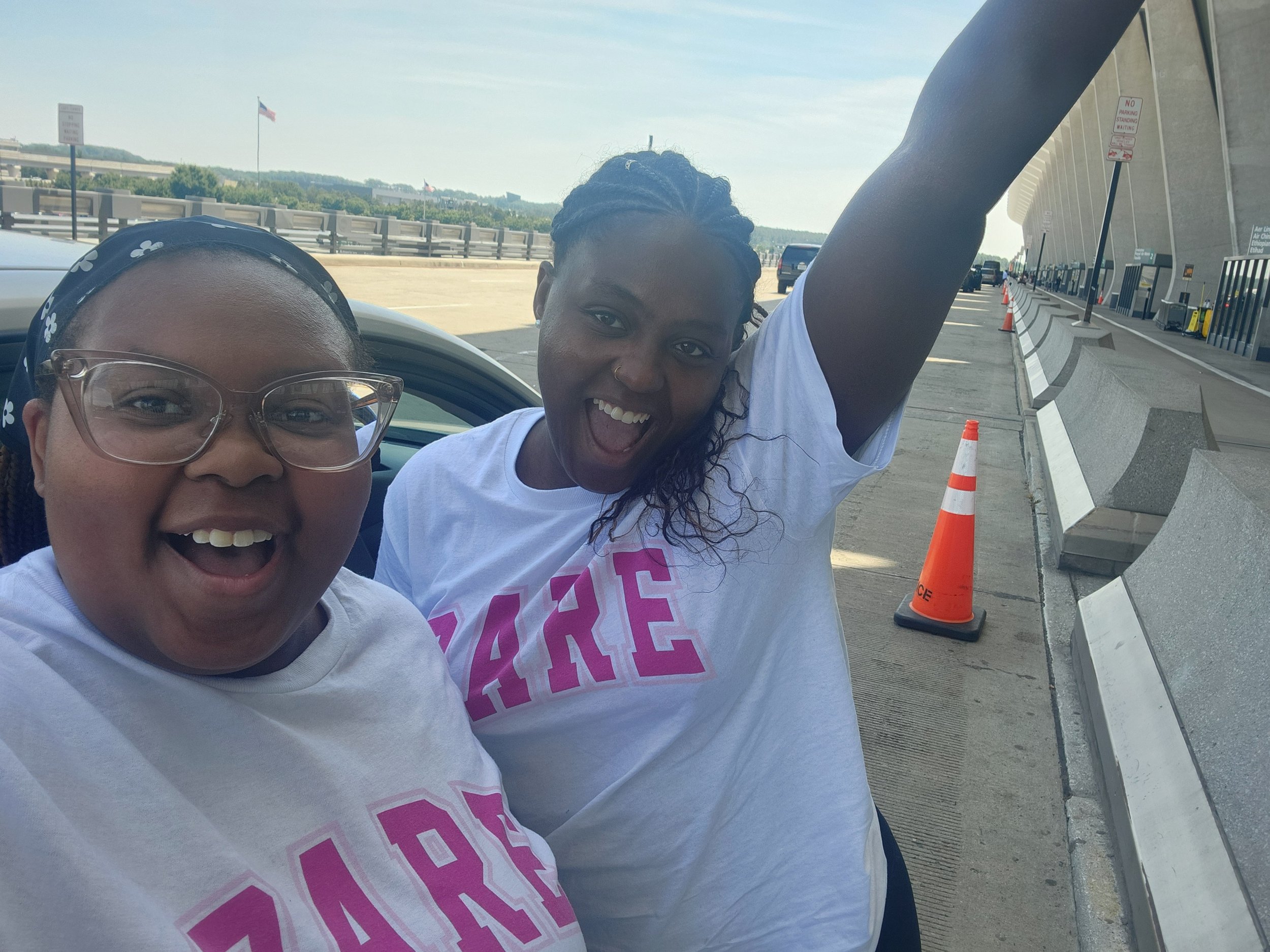 Two women smiling and appearing joyful outdoors on a bright day, standing next to a car with an urban background and orange traffic cones.
