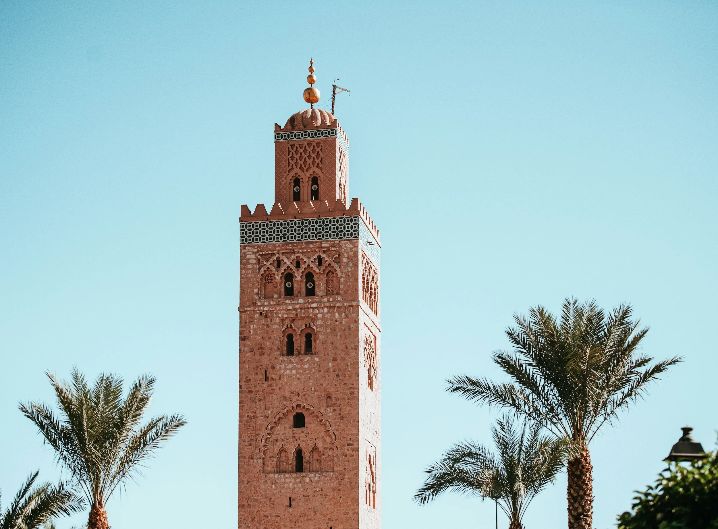 Tall red-brick tower with intricate Islamic architectural details, topped with a dome and golden ornaments, flanked by palm trees against a blue sky.