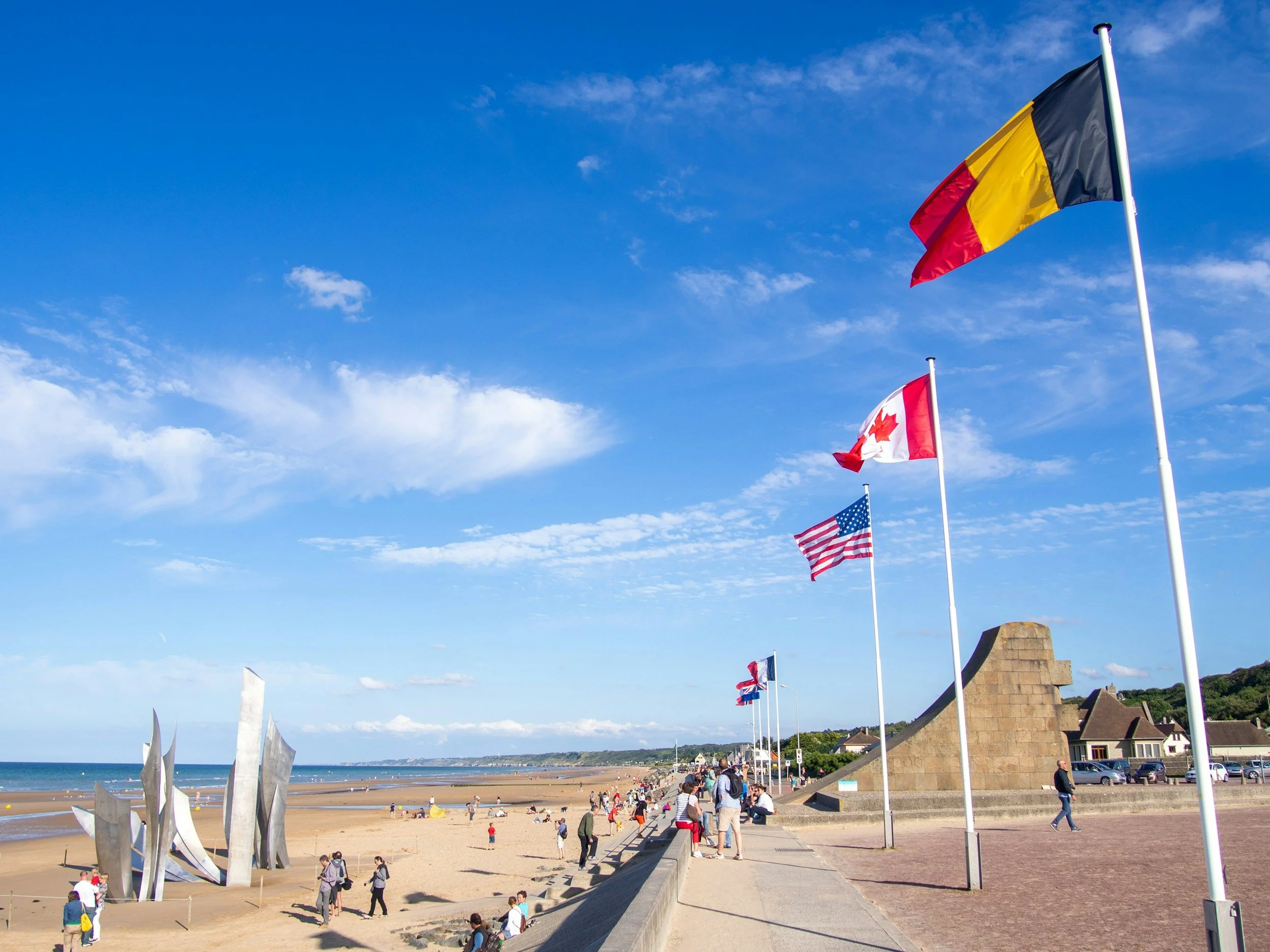 Beach with people, flags of Belgium, Canada, the United States, and the United Kingdom flying on flagpoles, and a modern sculpture on the sand, under a bright blue sky with some clouds.
